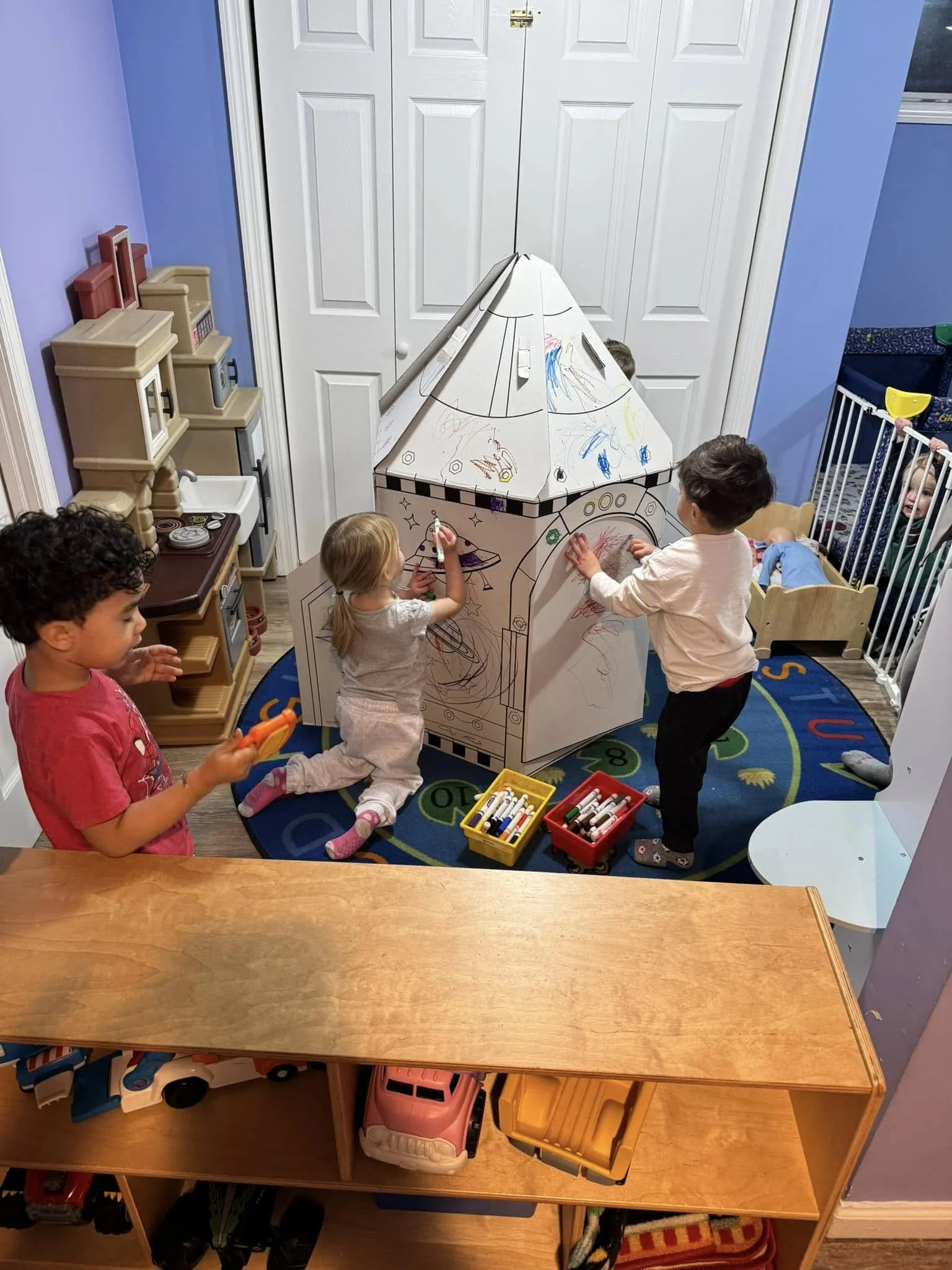 Children playing with a large cardboard spaceship decorated with markers in a colorful playroom.