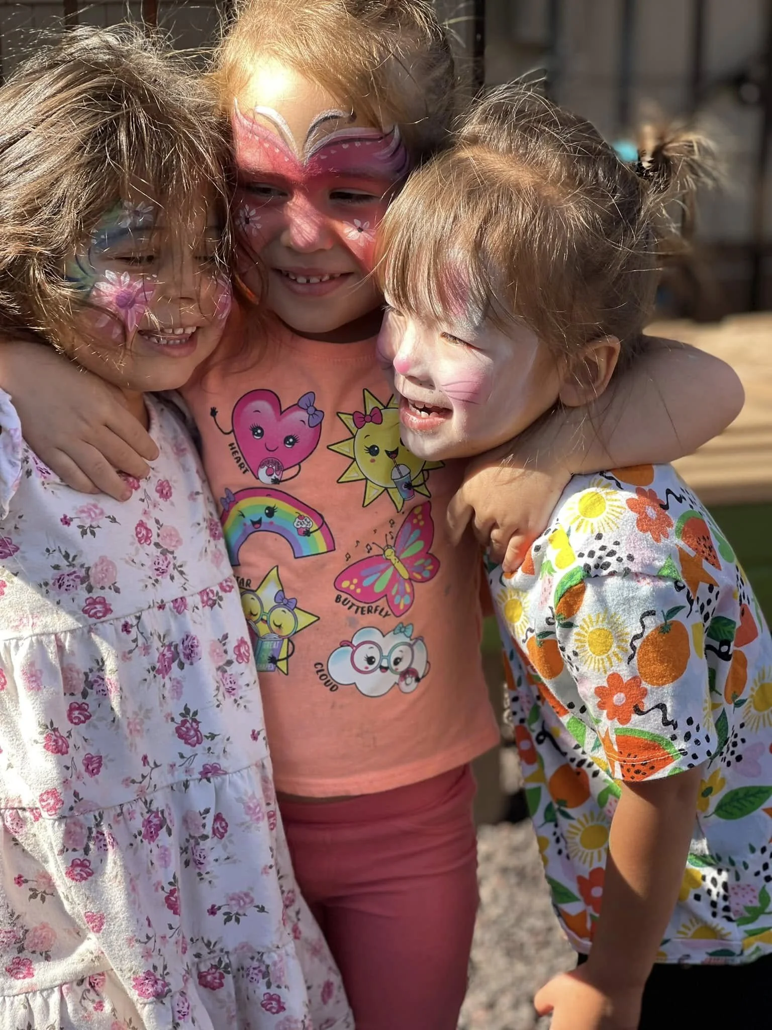Three young girls with face paint and colorful clothing hugging and smiling outdoors on a sunny day.