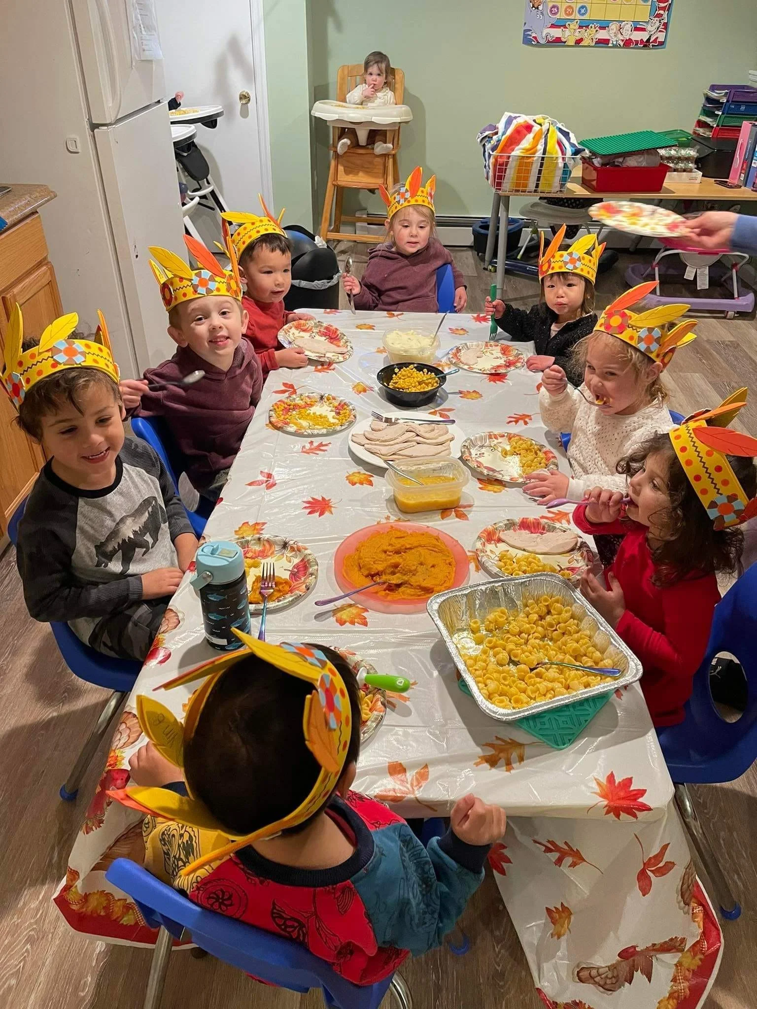 Children sitting around a table wearing colorful paper crown hats, celebrating with food including pasta, turkey slices, mashed potatoes, corn, and pumpkin dip.