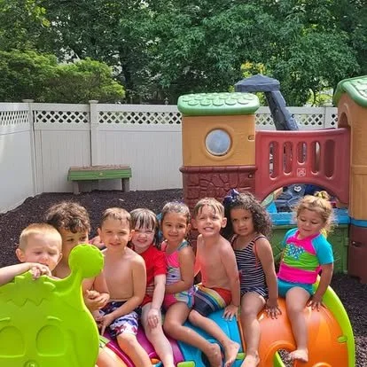 Children sitting on a colorful plastic water slide in a backyard with a white privacy fence and green trees.