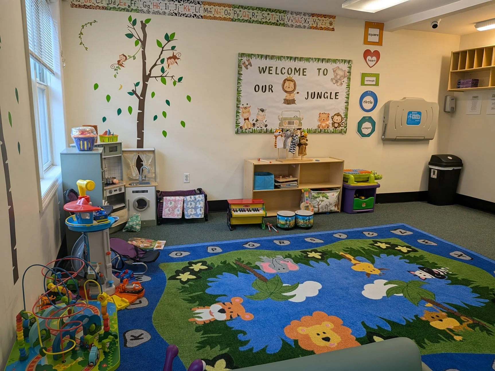 Colorful preschool classroom with jungle-themed decor, including a large jungle animal rug, wall decorations with animals and shapes, and shelves with toys and books.