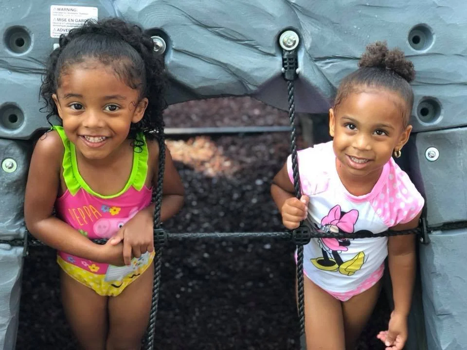 Two young girls smiling and peeking out of a play structure at a playground.