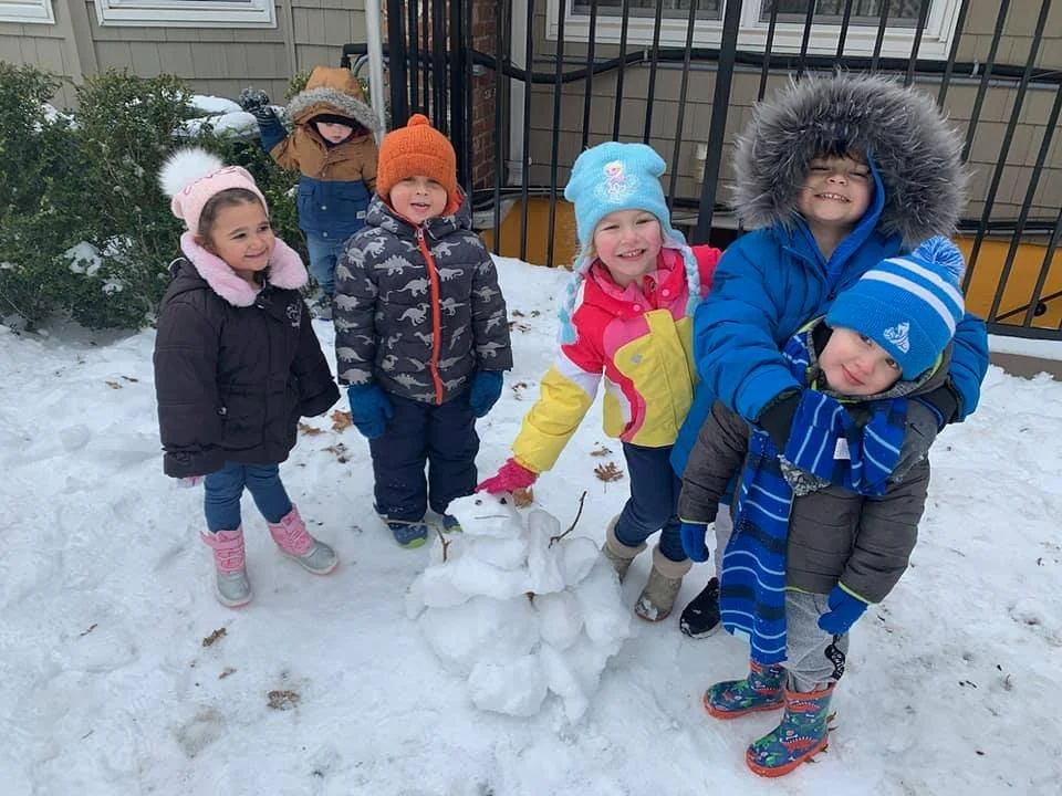 Group of six children in winter clothing outside near a snow-covered ground, standing around a small snowman they built with a stick for an arm, in front of a fence and some bushes.