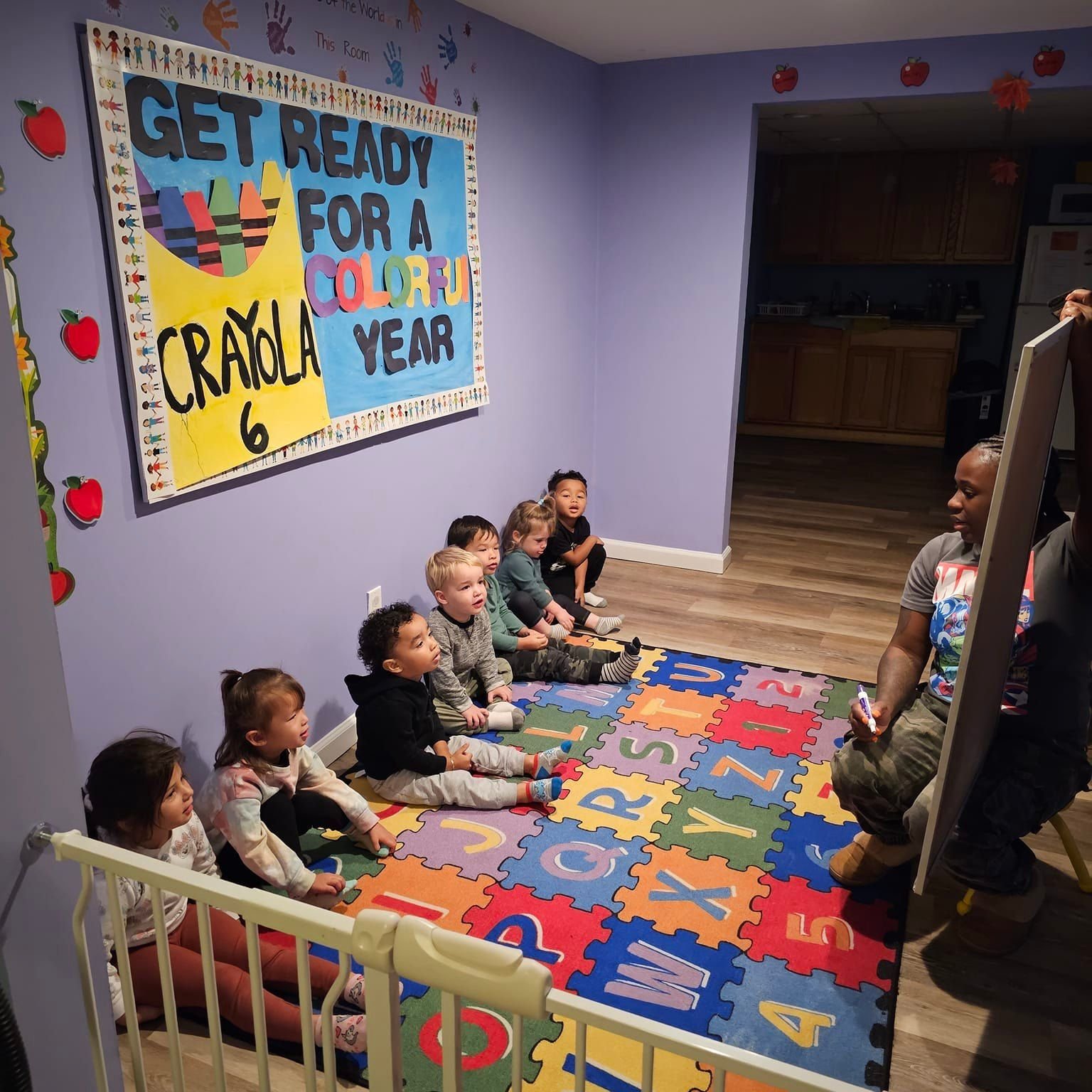 A group of young children sitting on a colorful alphabet rug in a classroom, listening to a woman sitting on a chair in front of them. The classroom walls are decorated with a large sign urging children to get ready for a colorful year, and there are apple and leaf cutouts on the walls. The setting is likely a preschool or kindergarten classroom.