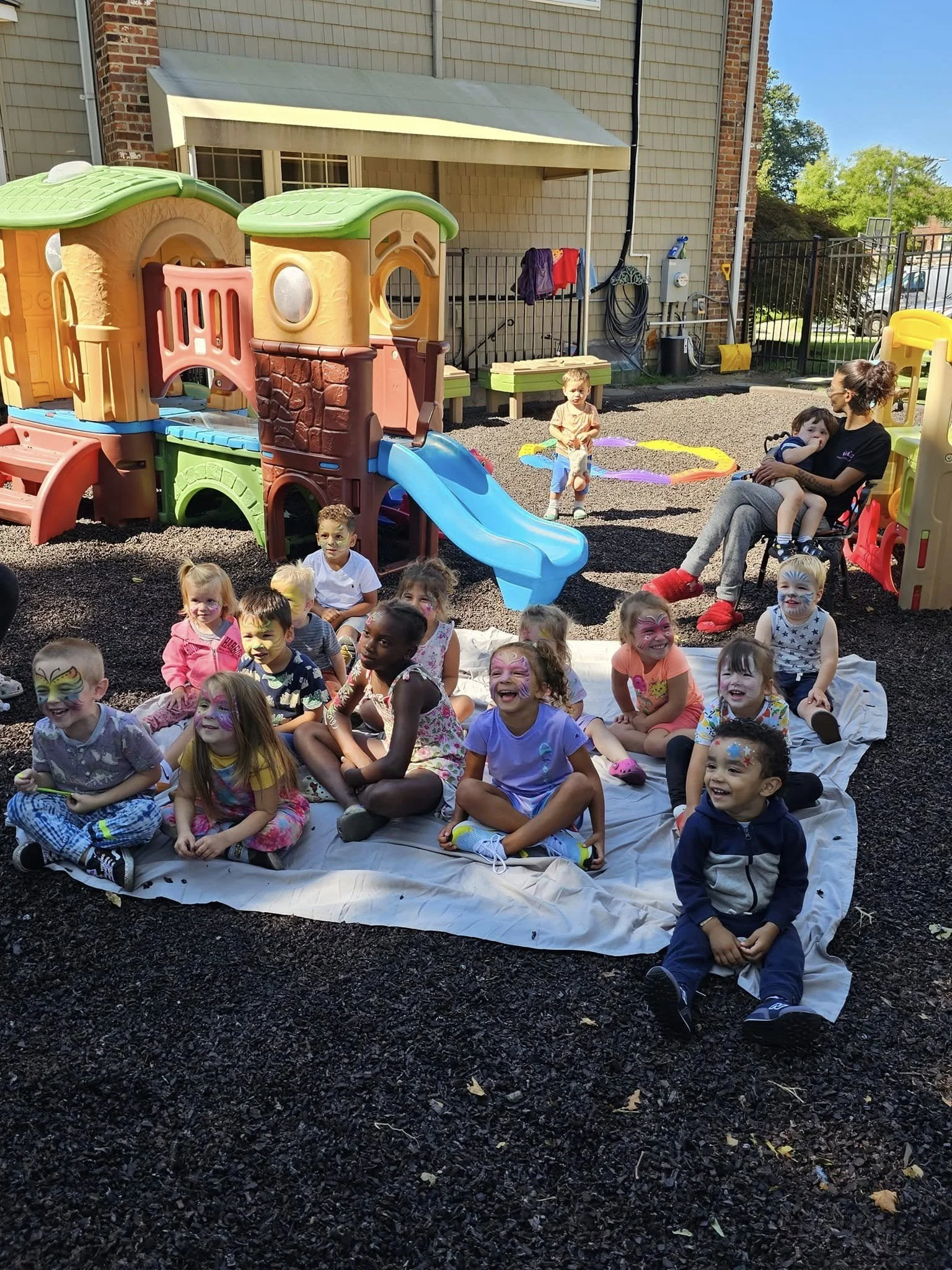 Group of young children with painted faces sitting on a white cloth on a playground, with a colorful plastic play structure and an adult supervising in the background.