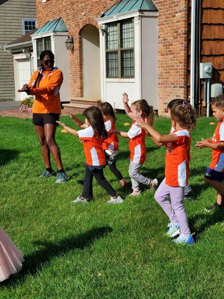 Children participating in outdoor activity with coach on green lawn in front of brick house.