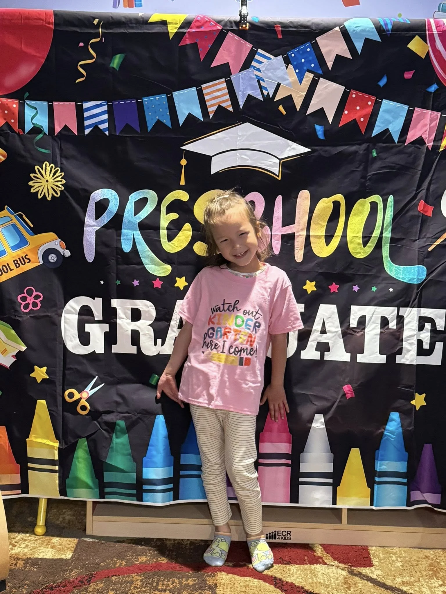 A young girl with a pink shirt and striped pants standing in front of a colorful graduation backdrop with the words 'Preschool Graduation' and various school-related illustrations, including a graduation cap, crayons, and a school bus.