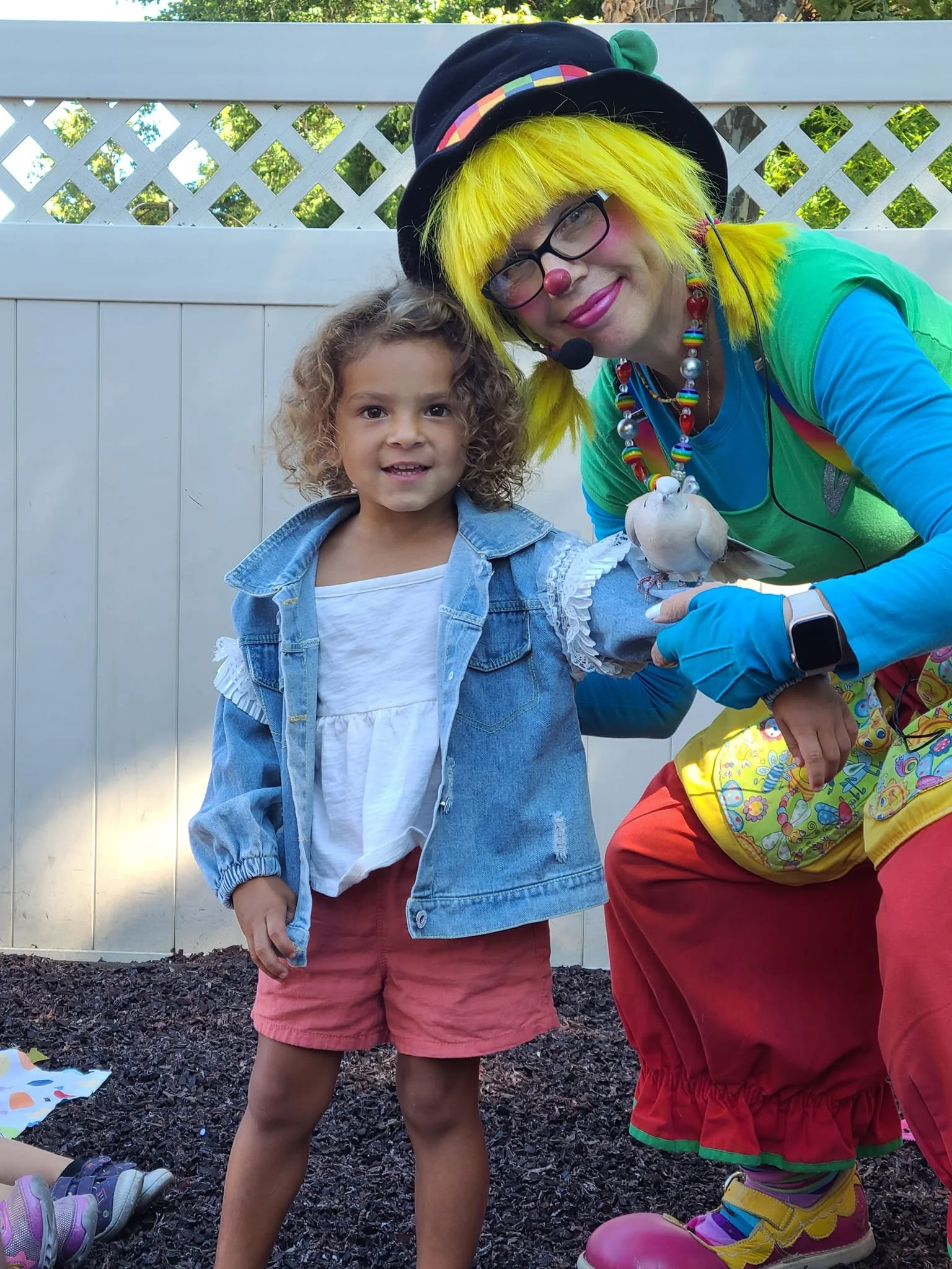 A young girl with curly hair standing next to a clown dressed in colorful costume, holding a white dove. They are outdoors in front of a white fence, with some toys visible on the ground.