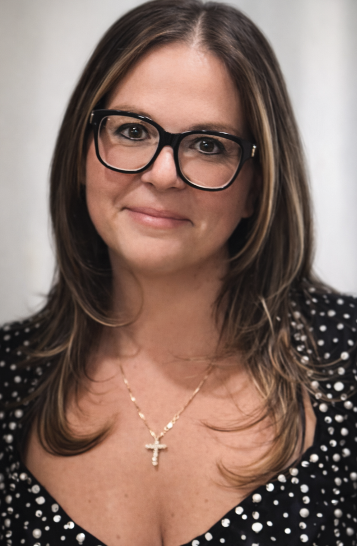A woman with brown hair wearing glasses, a black polka dot top, and a gold cross necklace.