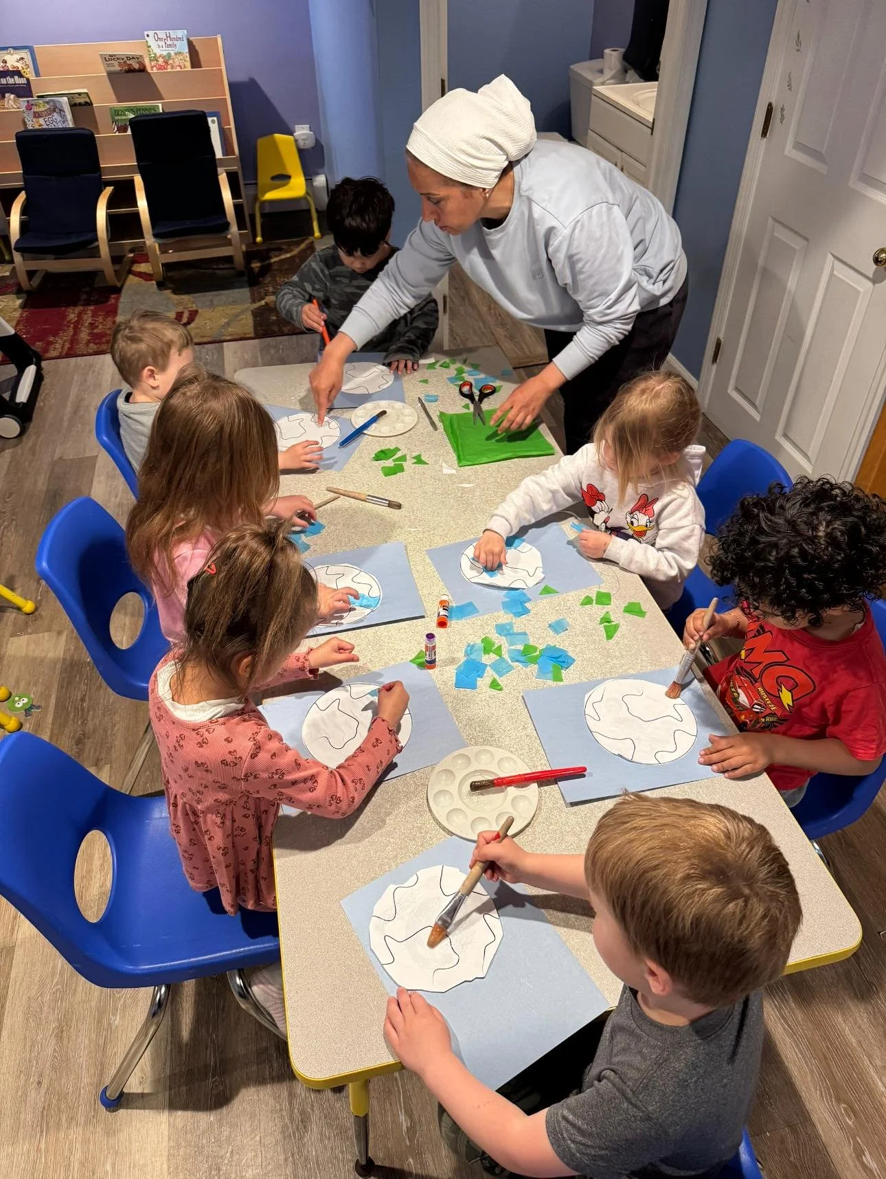 Children at a table doing Earth Day craft activity with teacher, gluing blue and green paper pieces onto Earth templates.