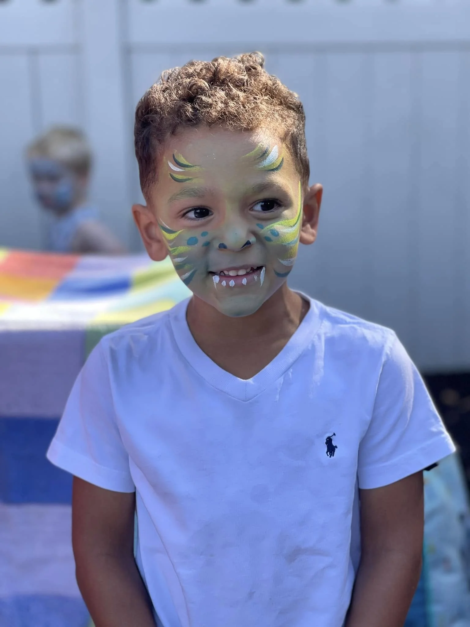 A young boy with face paint resembling a dragon, standing outdoors. He is wearing a white Ralph Lauren t-shirt, and another child with face paint is in the background.