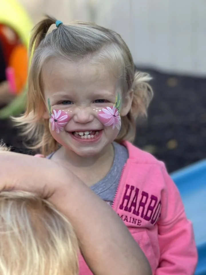 A young girl with face paint of pink flowers on her cheeks, smiling and showing missing teeth, wearing a gray and pink hoodie.