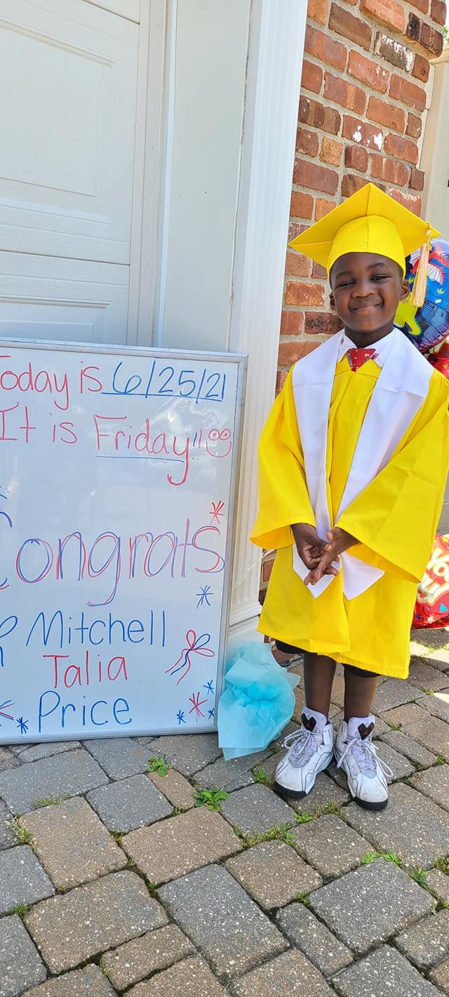 A young boy dressed in a graduation cap and gown standing next to a whiteboard with celebratory messages. The board says the date 6/25/21, that it's Friday, and congratulates Mitchell, Talia, and Price. The boy smiles and stands on a brick patio in front of a brick wall and a garage door.