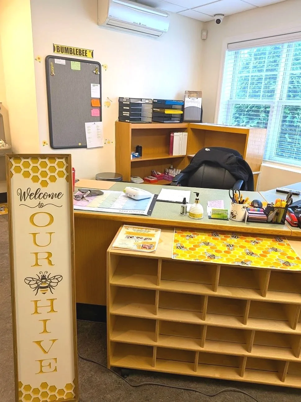 Office reception desk decorated with a bee and honeycomb theme, featuring a welcome sign, small cubby shelves, and various office supplies. There is a large window with blinds, and shelves behind the desk contain files, organizers, and office equipment.