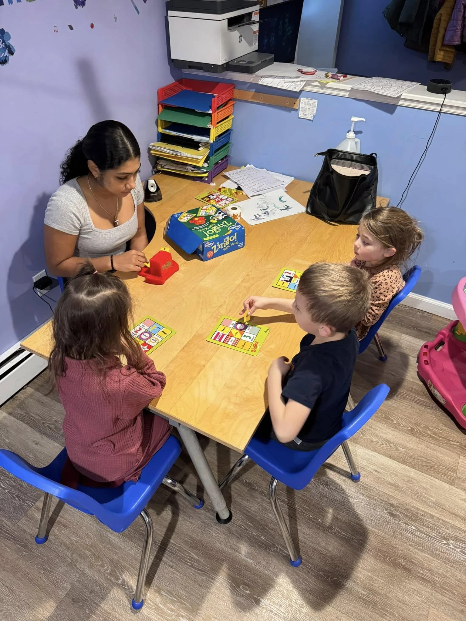 A teacher and three children playing a board game at a table in a classroom.