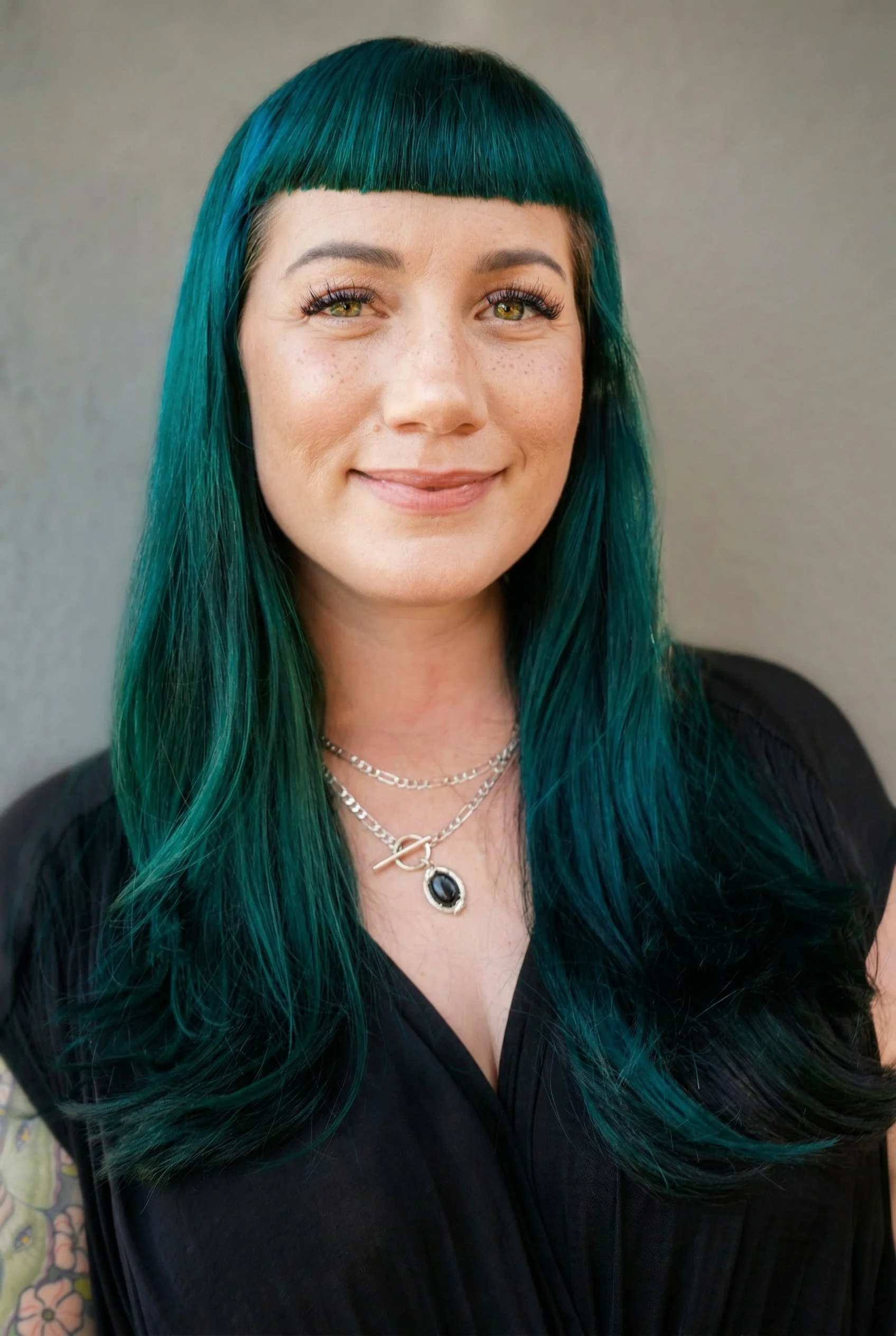 Close-up of a woman with shoulder-length teal hair, bangs, and hazel eyes, smiling and wearing layered silver necklaces and a black top.