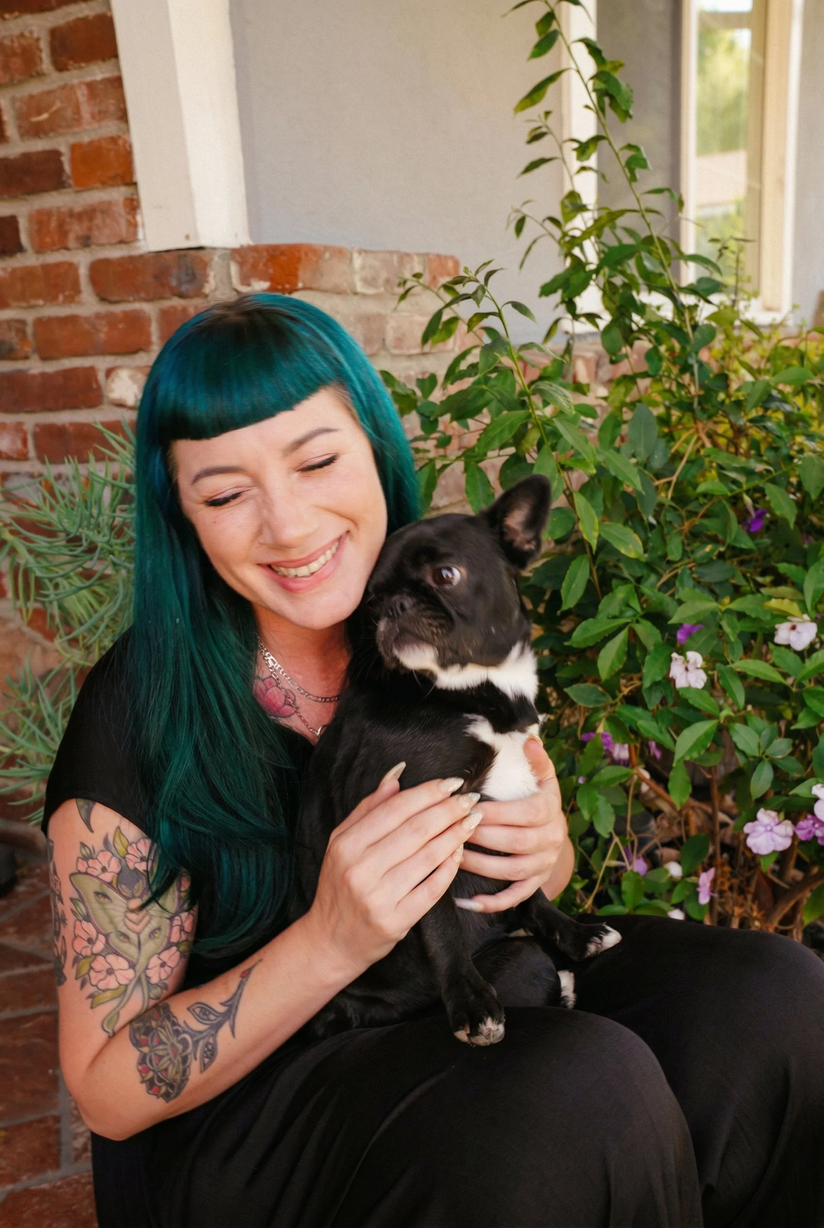 Woman with teal hair smiling while holding a black and white French Bulldog puppy surrounded by green plants and a brick wall.