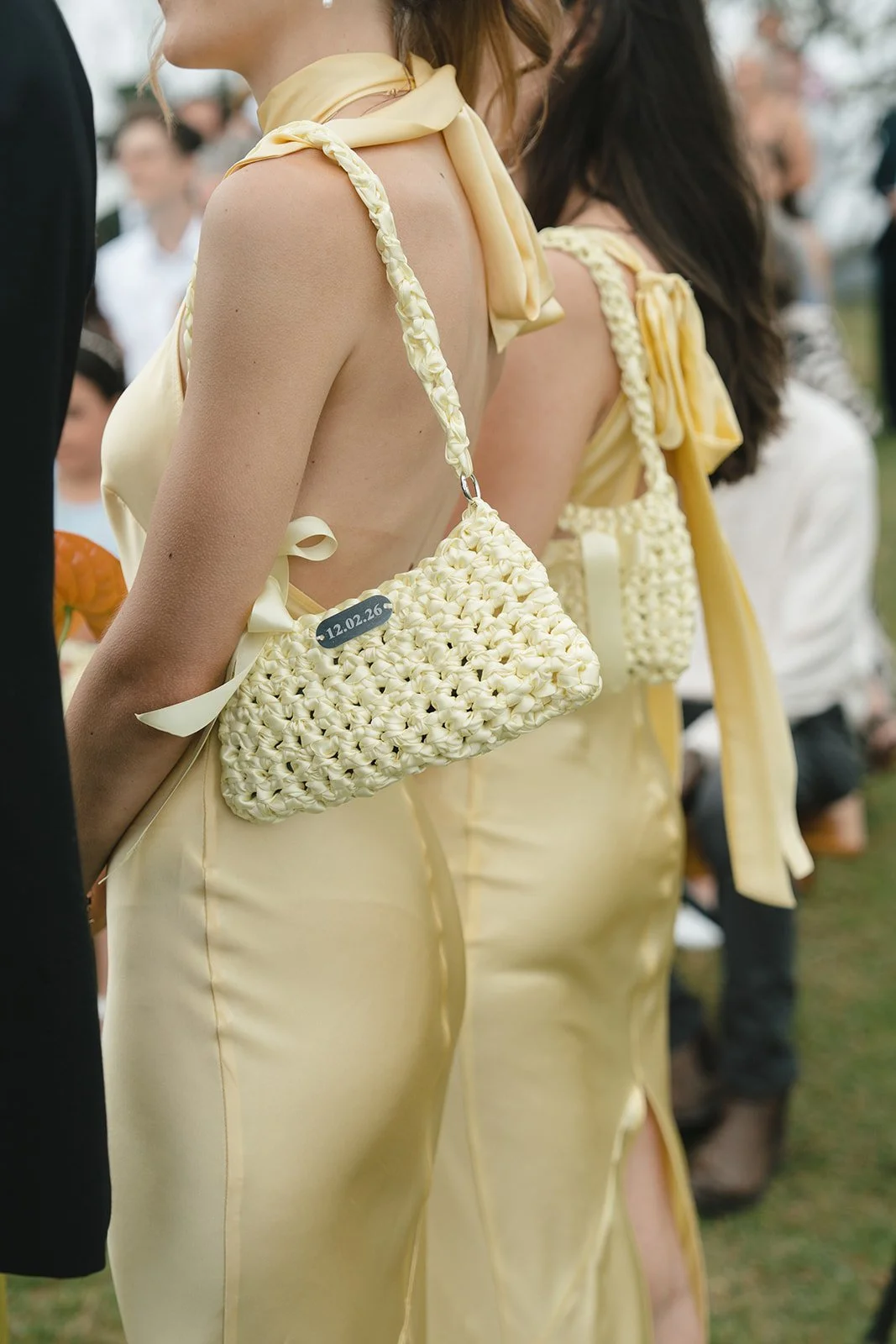 Close-up of women in yellow satin dresses with braided handbags at an outdoor event, with people in the background.