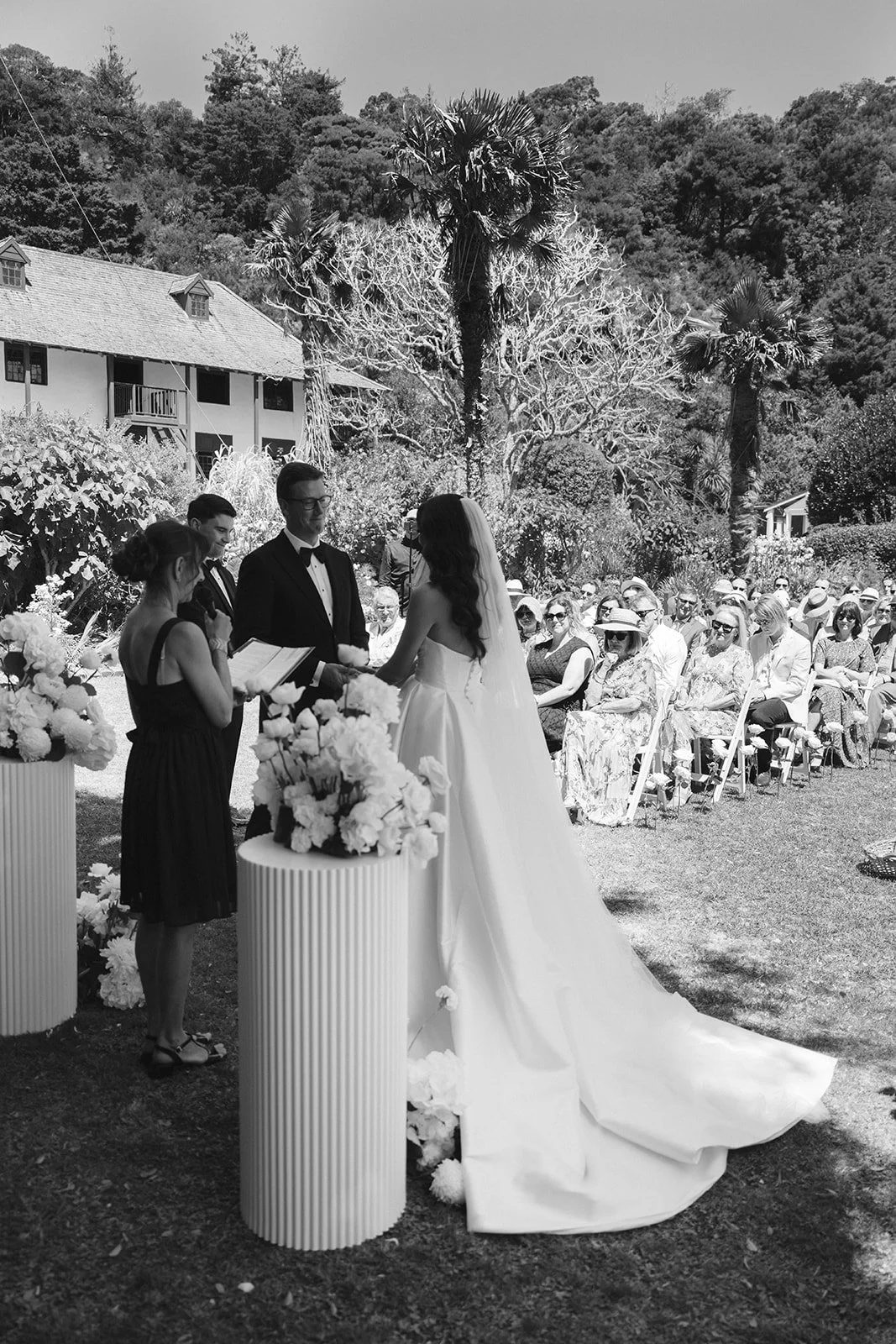 Black and white photo of a wedding ceremony outdoors with a bride and groom exchanging vows, surrounded by guests seated in chairs, with trees and a house in the background.
