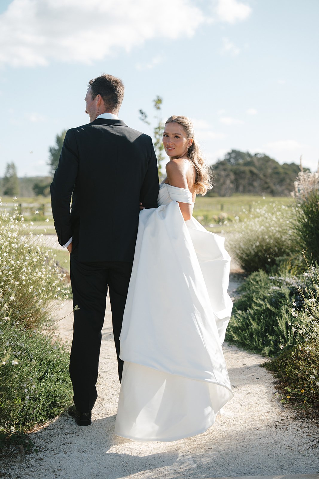 A bride and groom standing outdoors on a sunny day, with the bride looking back over her shoulder. The bride wears a white off-shoulder gown, and the groom is dressed in a black suit. They are surrounded by greenery and flowers.