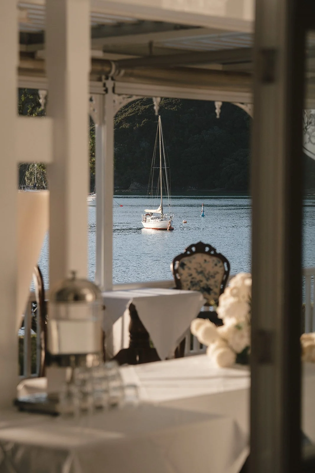 A view through a window showing a sailboat on calm water with a forested hill in the background, framed by a decorated dining setup in the foreground.