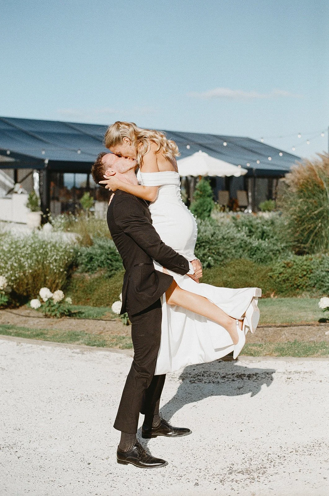 A man in a black suit lifting a woman in a white wedding dress outside on a sunny day, with greenery and a building in the background.