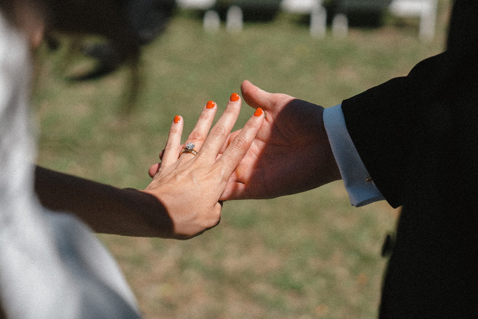 Close-up of a couple holding hands during a wedding or engagement ceremony. The woman has a diamond ring on her ring finger and orange nail polish. The man is wearing a black suit with a light blue shirt. The background is outdoors with blurred grass and white chairs.
