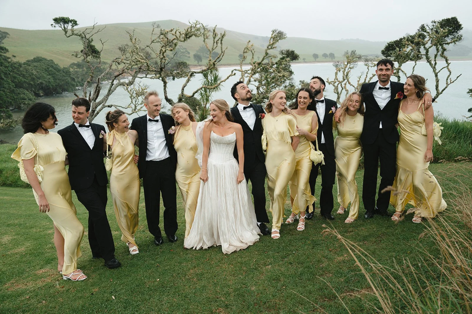 A wedding party of fifteen people, including the bride in a white gown and fifteen bridesmaids and groomsmen in formal attire, standing outdoors on grass near trees with a lake and hills in the background, smiling and laughing.