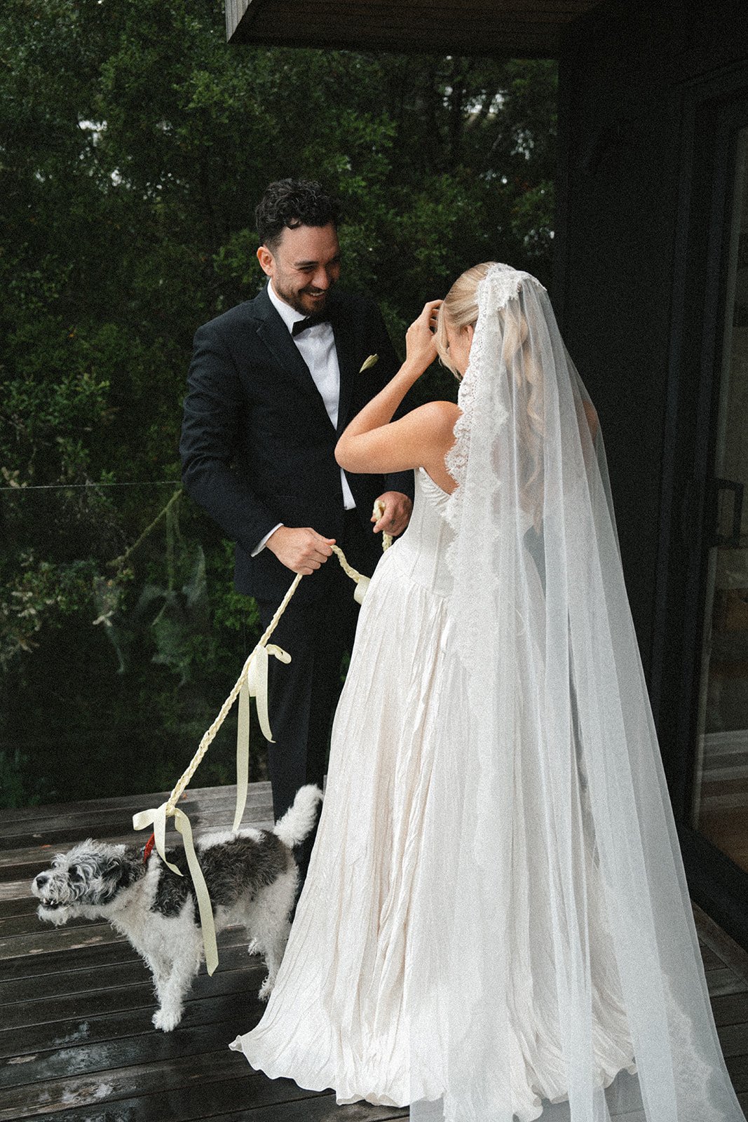 A bride and groom on their wedding day, standing on a wooden deck, with the groom holding a leash attached to a small black and white dog. The bride is wearing a white wedding dress and veil, while the groom is dressed in a black suit with a bow tie. They are smiling and looking at each other, with lush greenery in the background.