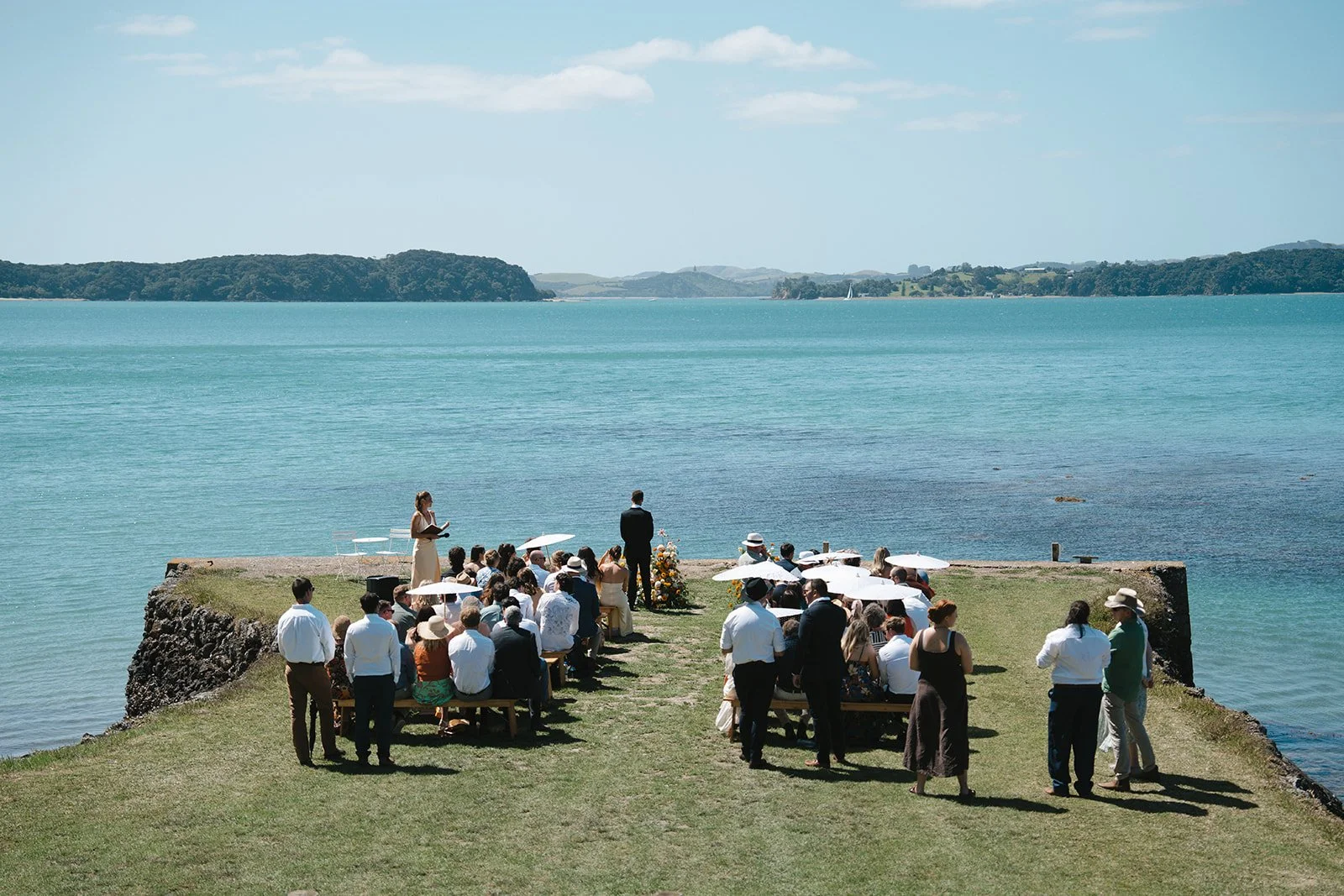 A wedding ceremony taking place on a grassy ledge overlooking the water, with guests seated and standing around a bride and groom, under a clear sky with calm blue water and green hills in the background.