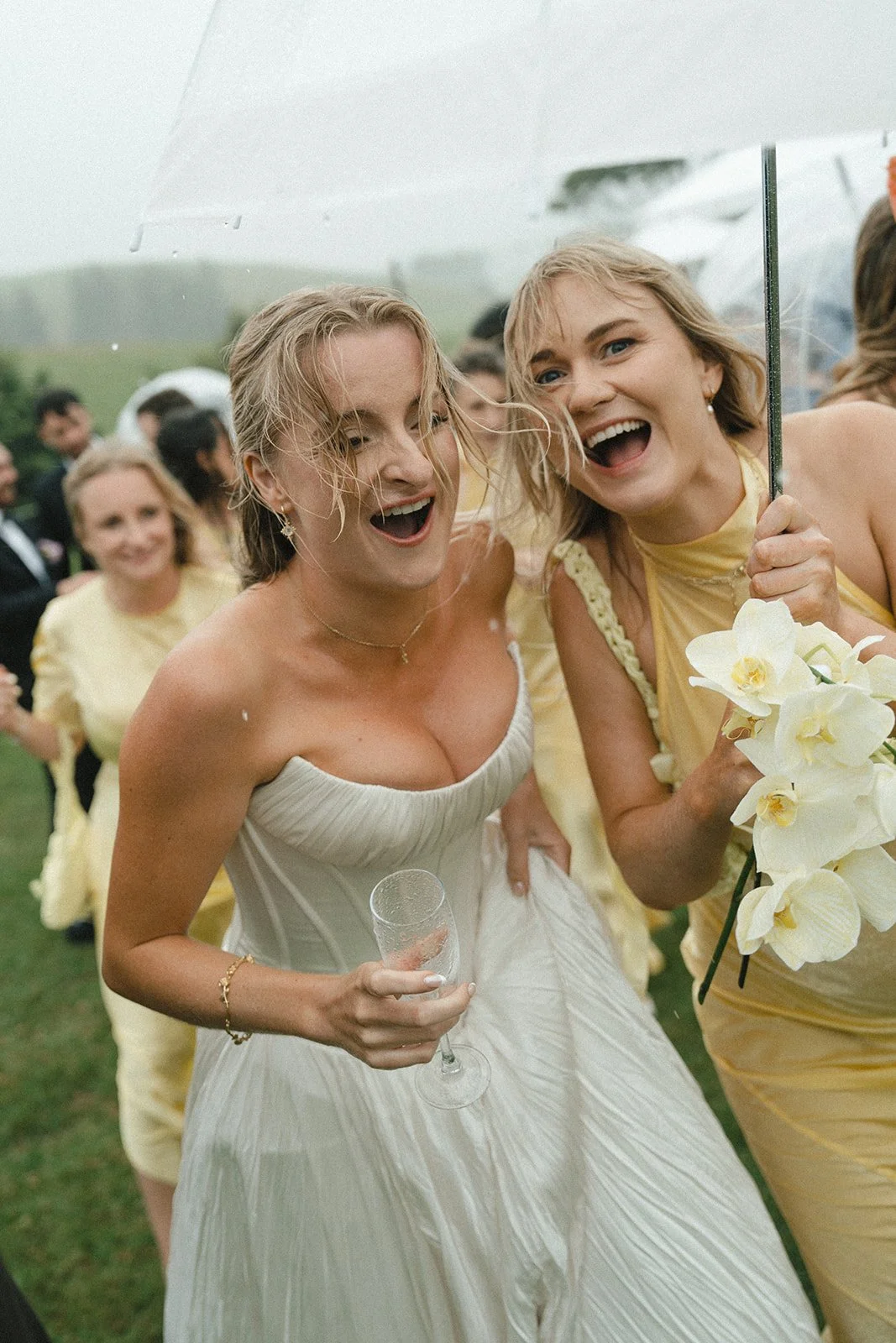 Two women celebrating outdoors on a rainy day, smiling, with others in the background, one in a wedding dress holding a glass and the other holding a bouquet of white flowers, everyone wearing light yellow outfits.