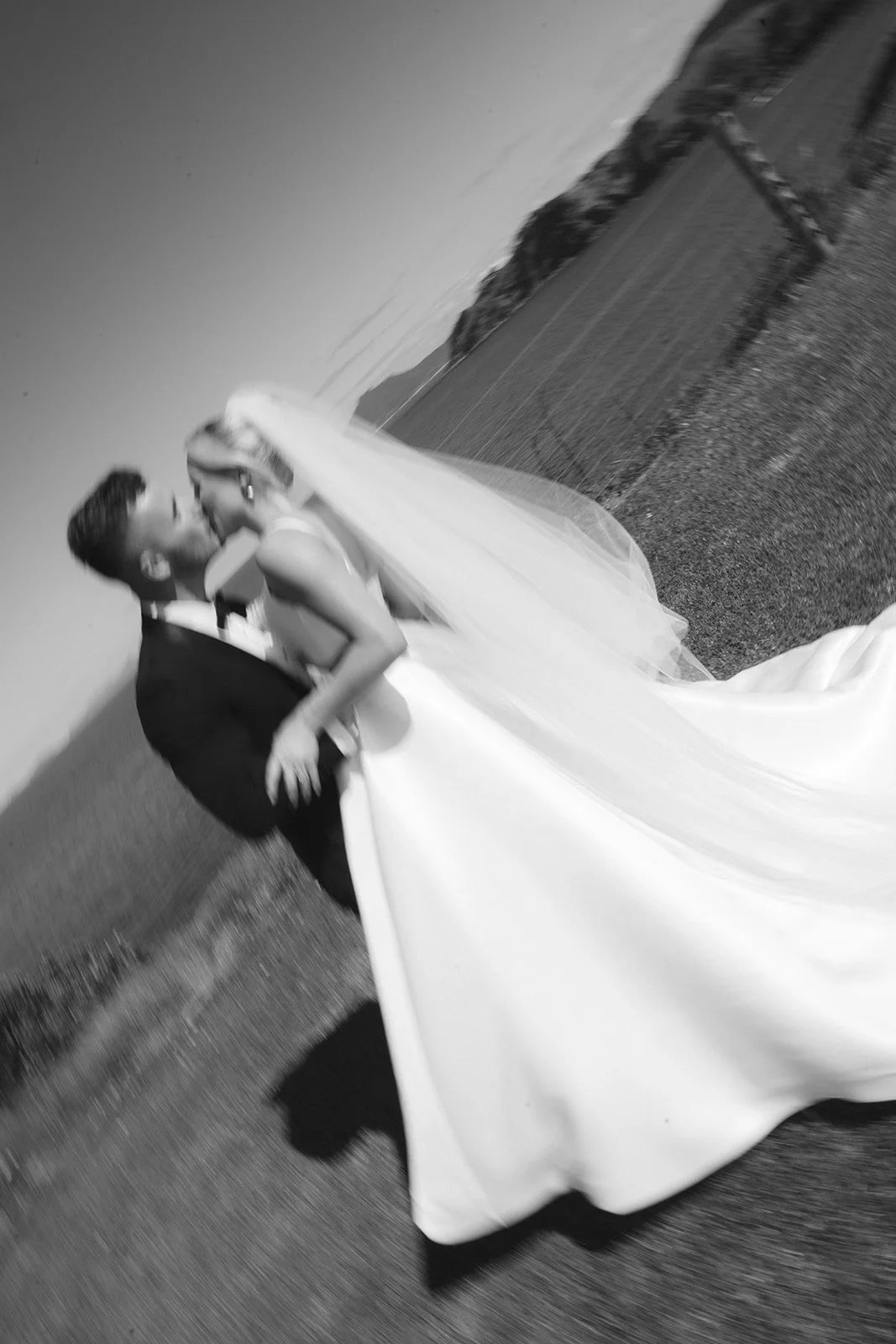 A couple in wedding attire sharing a kiss outdoors, with the bride in a white gown and veil and the groom in a suit, against a background of field and hills.