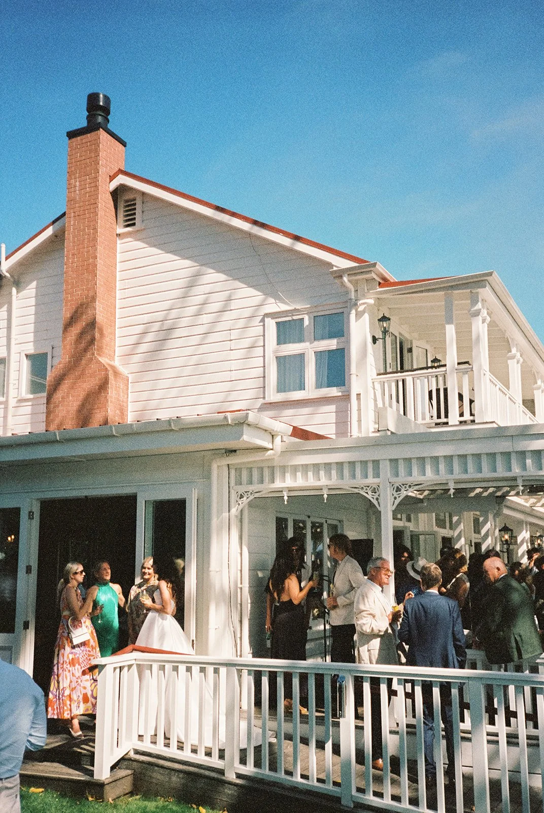 People socializing outside a white house with a porch and a red brick chimney under a clear blue sky.
