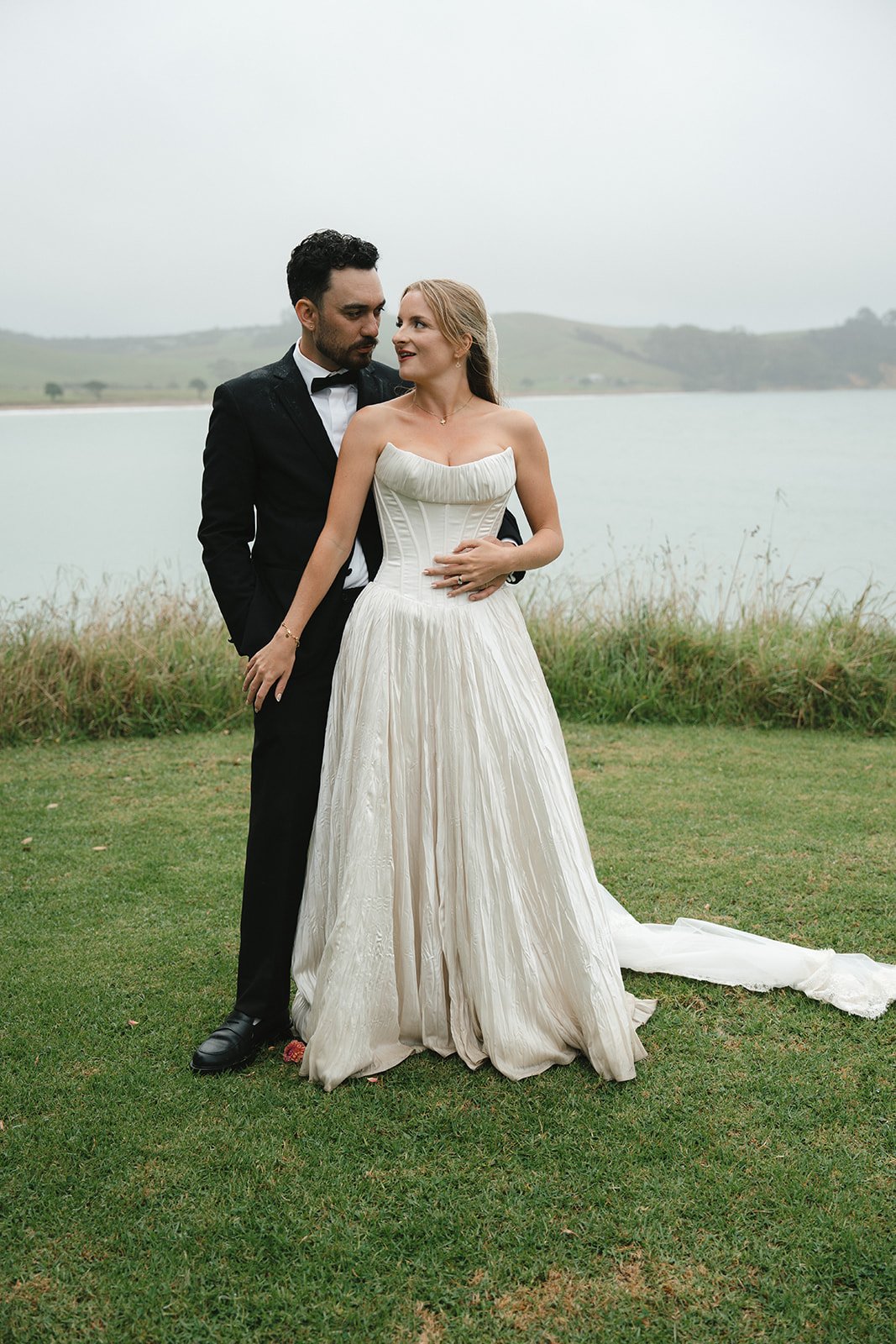 A bride and groom standing outdoors on grass near a lake, with the groom in a black tuxedo and the bride in a strapless white wedding gown, sharing a tender moment.