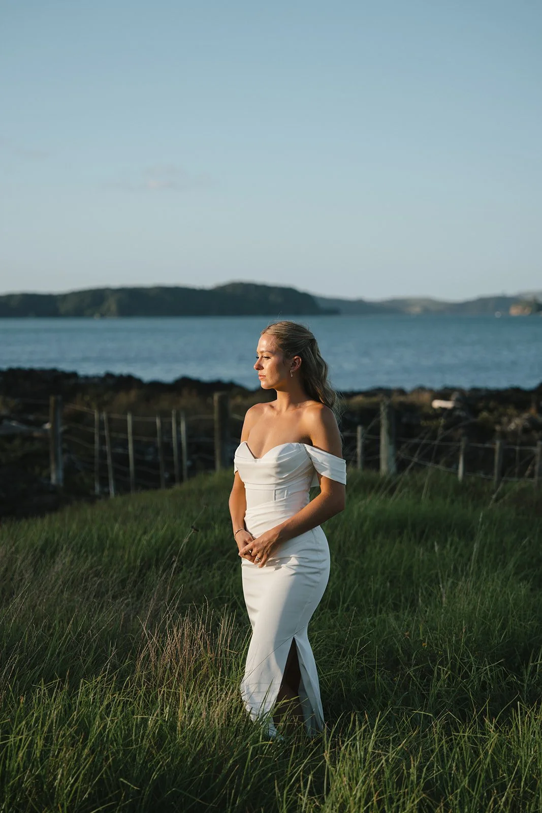 A woman in a white off-the-shoulder dress standing in grassy field near water with hills in background.