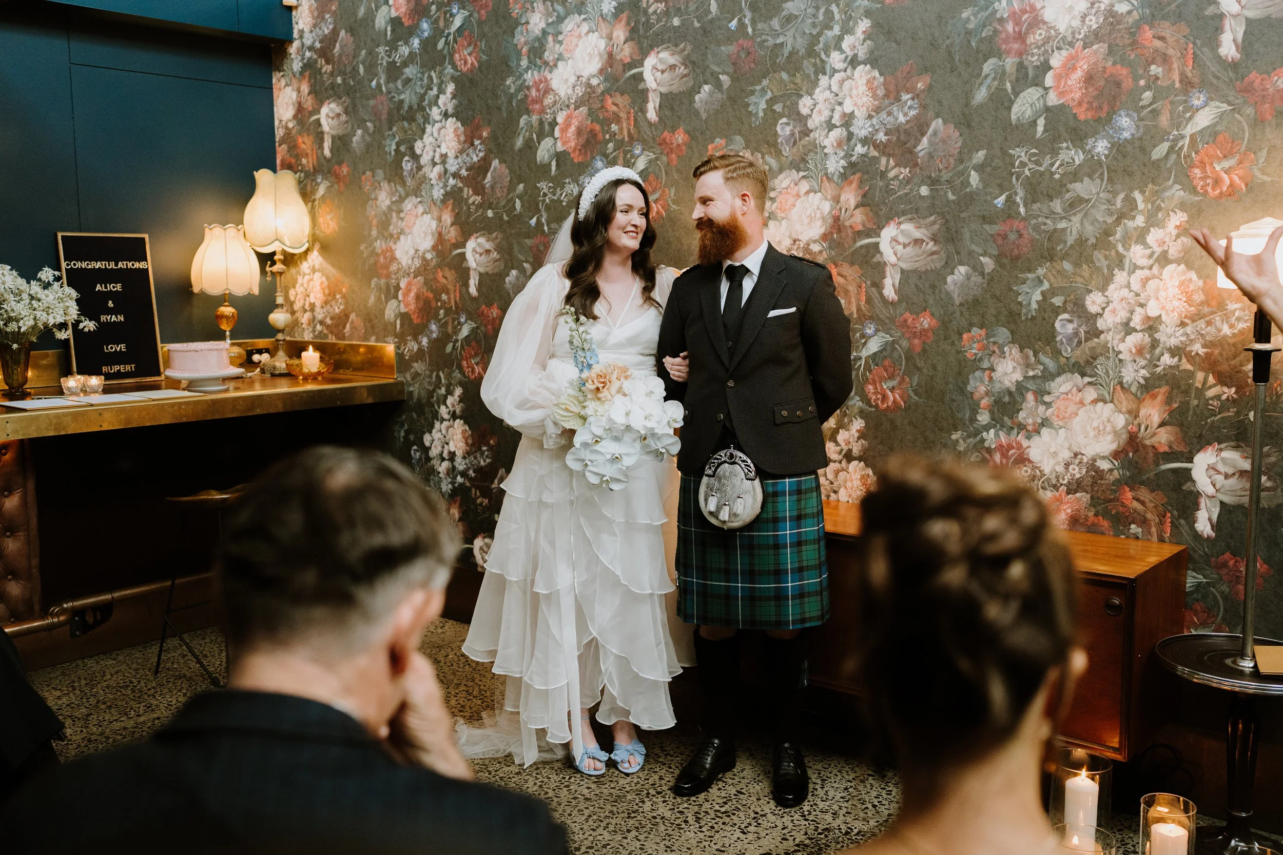 A bride and groom standing close together and smiling during their wedding ceremony. The bride is wearing a white gown and holding a bouquet of flowers, while the groom is wearing a black suit and a traditional Scottish kilt. They are inside a room with a floral wallpaper background, with guests sitting in front of them.