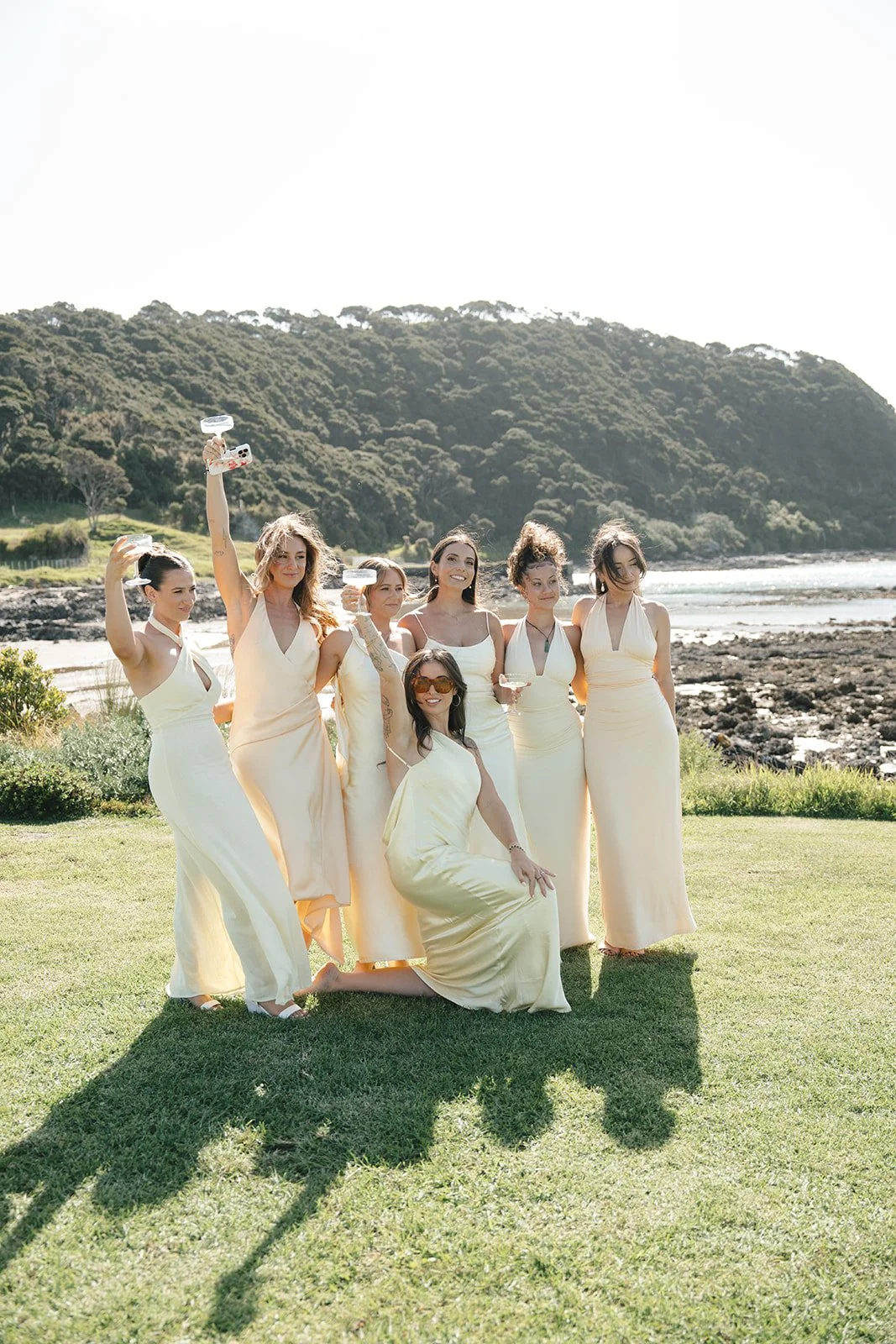 Group of women in cream-colored dresses celebrating outdoors near a beach, holding drinks, with a green hillside and rocky shoreline in the background.