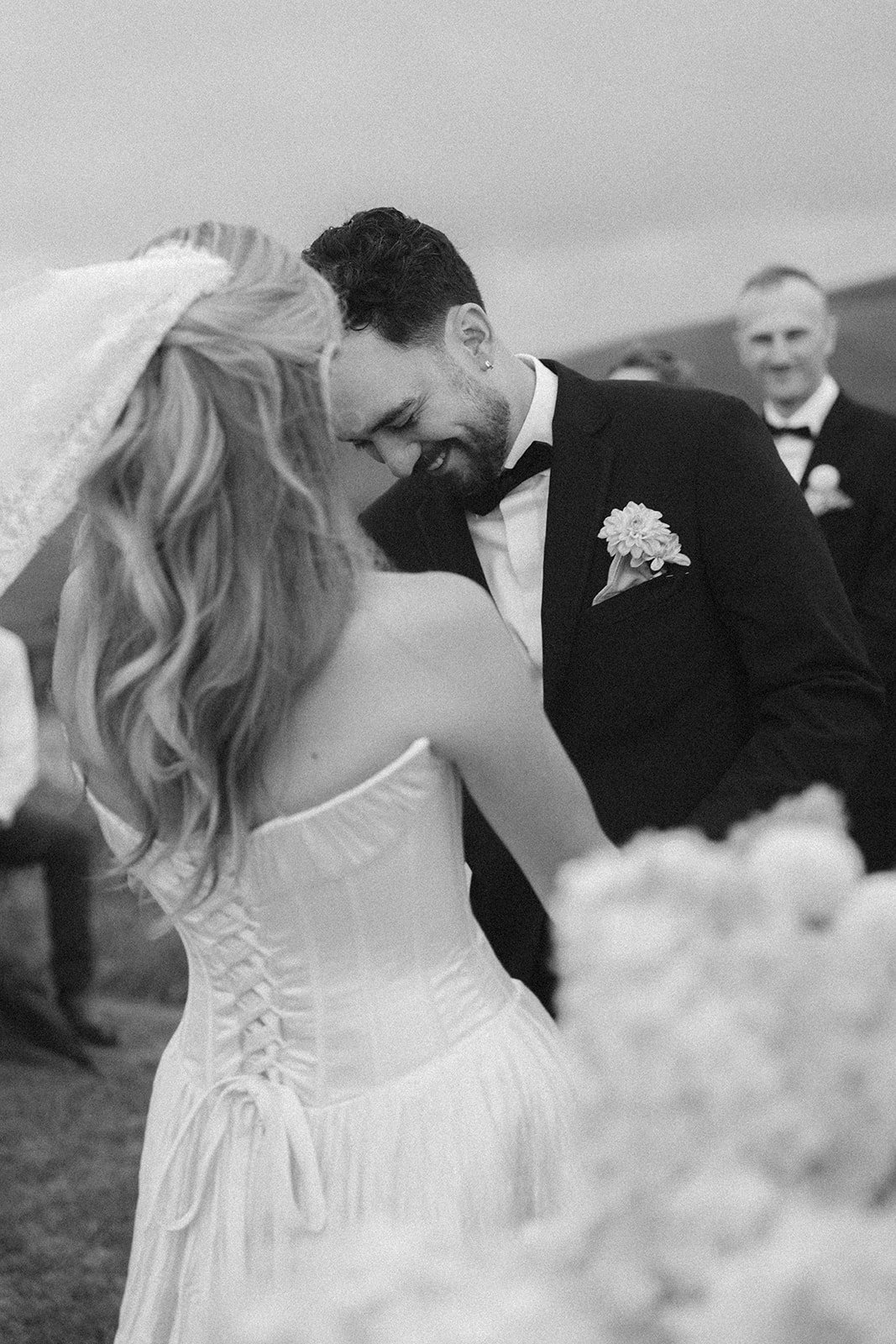 A black and white photo of a wedding scene showing a bride and groom sharing a joyful moment, with the groom smiling and the bride with long hair and a strapless gown. There are wedding guests in formal attire in the background.