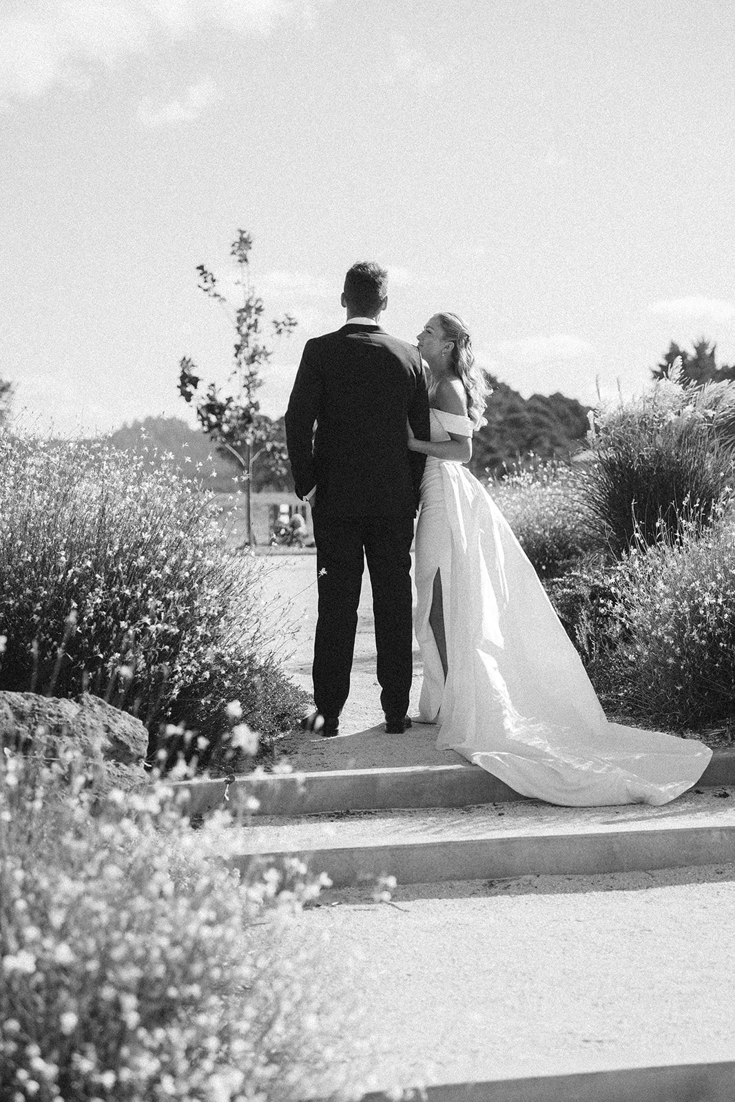 A black and white photo of a bride and groom walking arm in arm outdoors on a sunny day, surrounded by tall grasses and trees, with the bride in a long flowing wedding gown and the groom in a suit.
