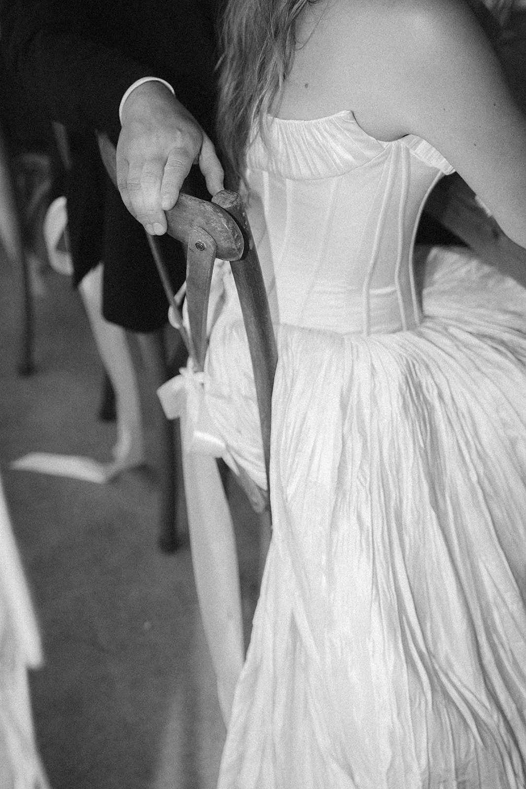 A person in a white dress sitting in a wheelchair, holding the wheelchair's wooden armrest, in a black-and-white photograph.