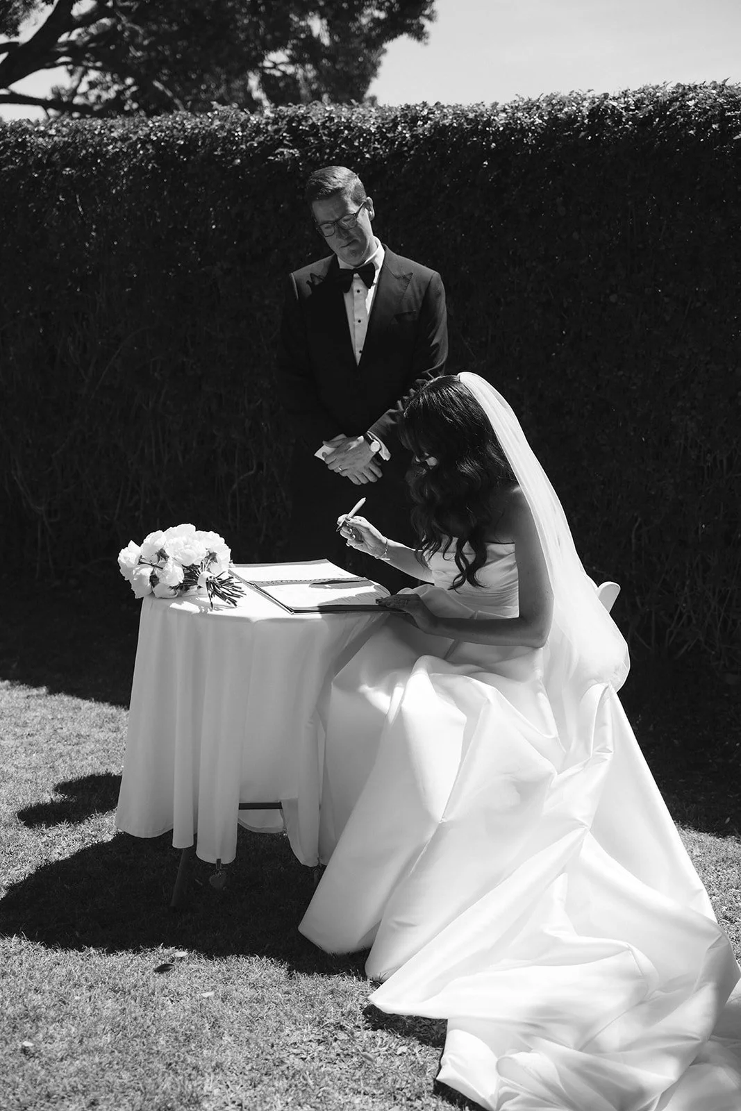 Black and white photo of a bride signing a marriage document while a groom stands beside her outdoors.