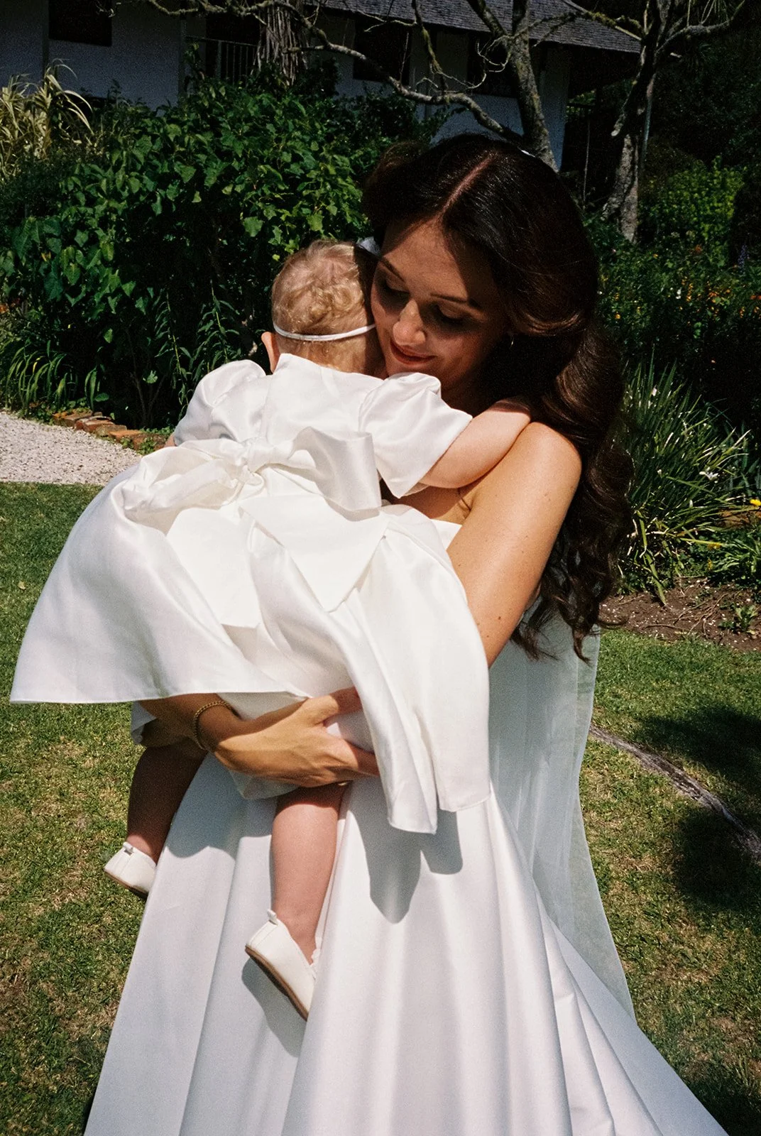 Woman in a wedding dress holding a young child in a white outfit, outdoors in a garden at night.
