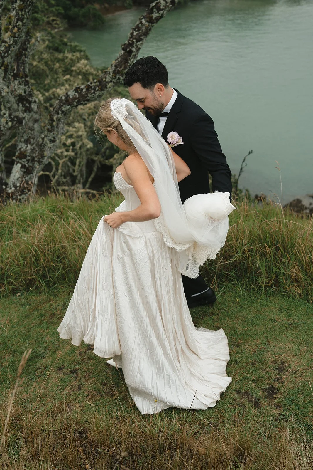 A bride and groom in wedding attire sharing a kiss outdoors near a river, with trees and grass around them.