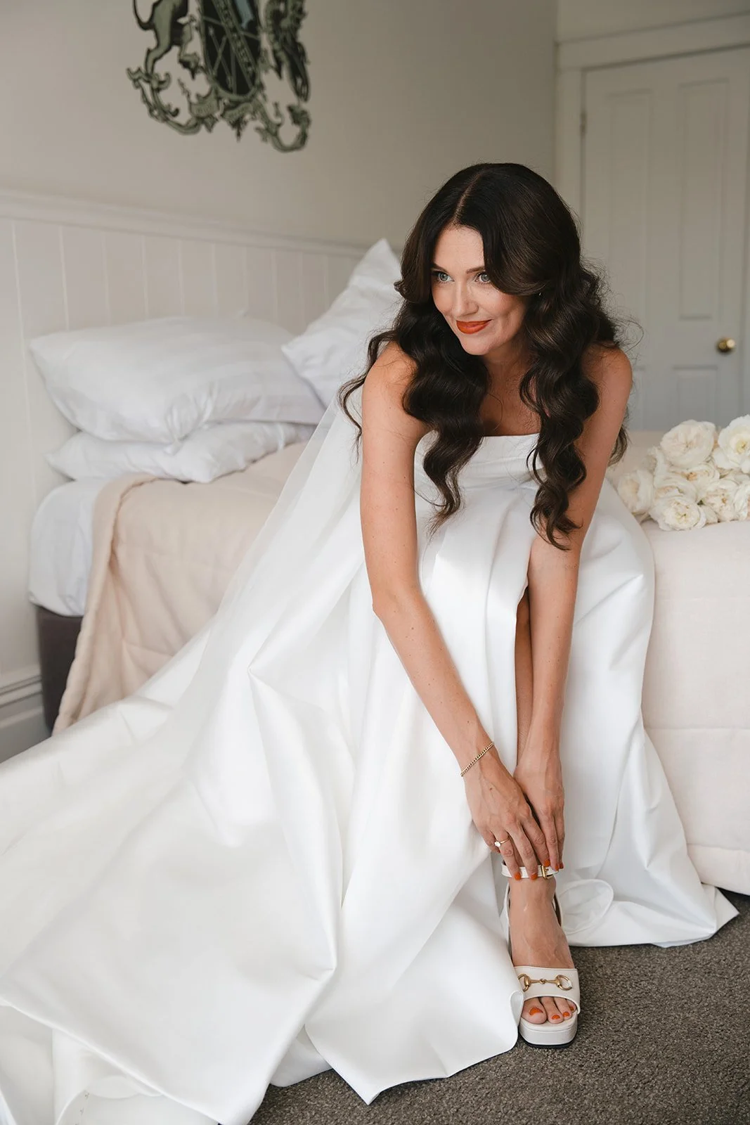 A woman in a wedding dress sitting on the edge of a bed, adjusting her shoe, with dark wavy hair and makeup in a cozy bedroom.