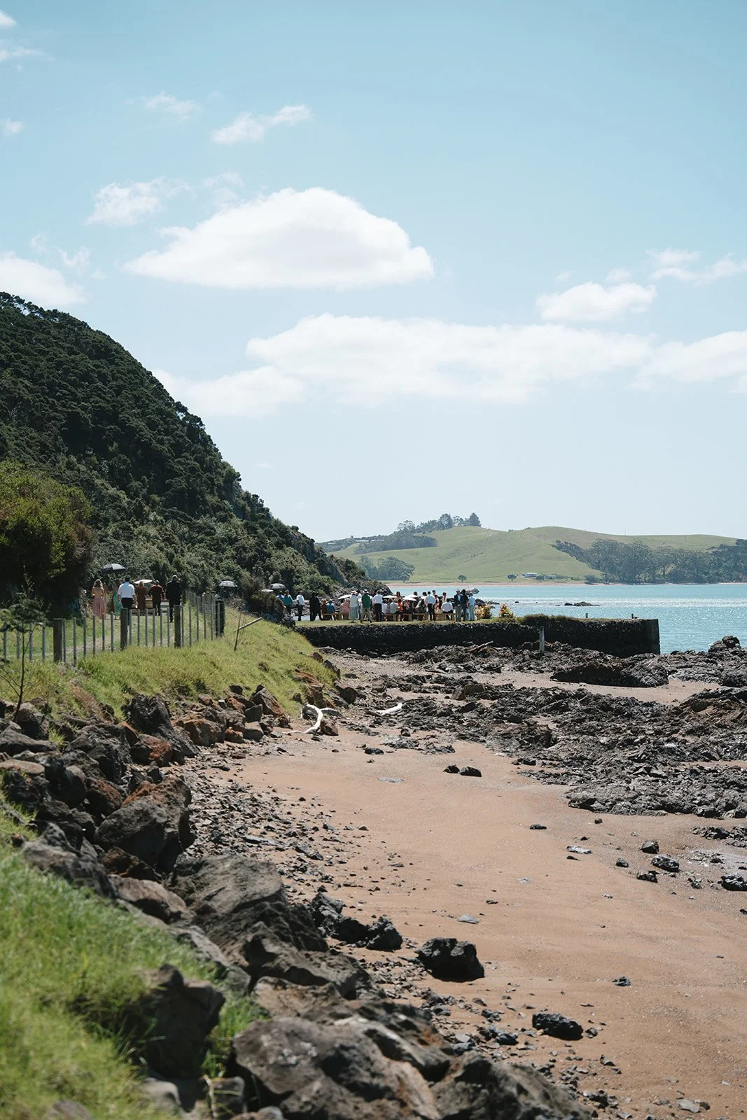 A coastal scene with a rocky beach in the foreground, a grassy hillside on the left, and a group of people standing near the water's edge under umbrellas. In the background, there are rolling green hills under a partly cloudy sky.