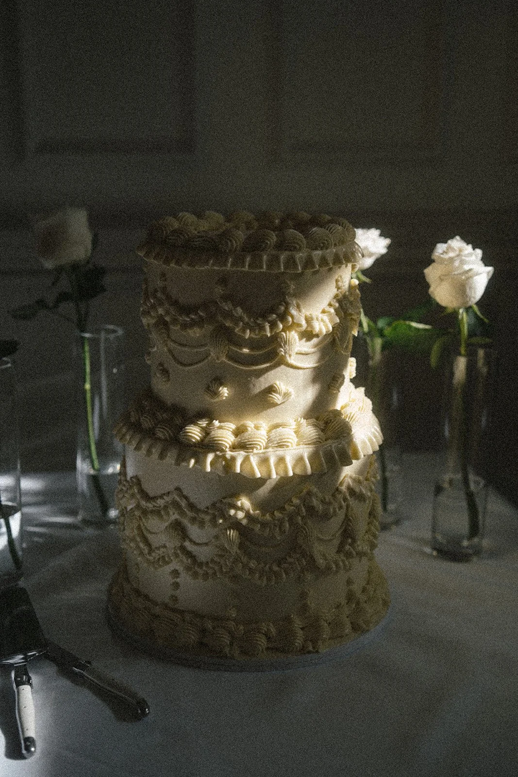 A multi-tiered decorative cake with intricate icing designs, surrounded by white roses in vases, in a dimly lit setting.