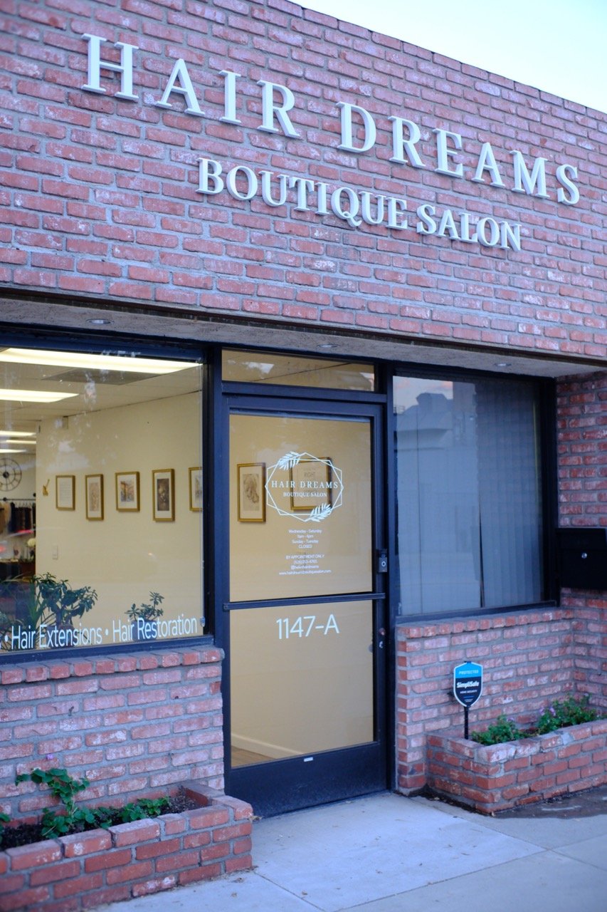 Exterior of a brick building with a sign reading 'Hair Dreams Boutique Salon' above the entrance. The door has the salon's logo and business hours, and there are planters with plants on either side of the door.