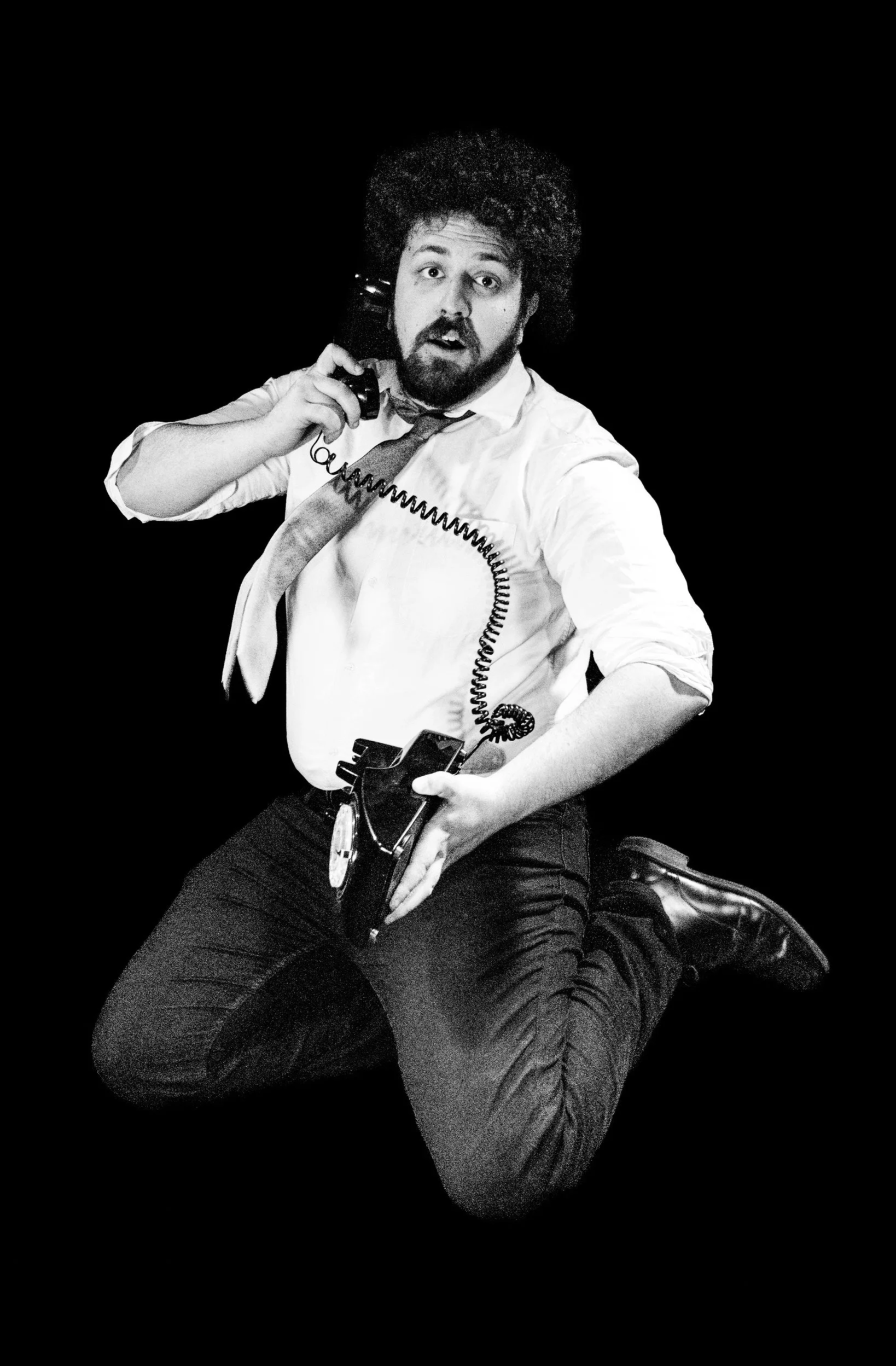 Black and white photo of a man sitting on a motorcycle, holding a telephone receiver with one hand and the motorcycle with the other, against a dark background.