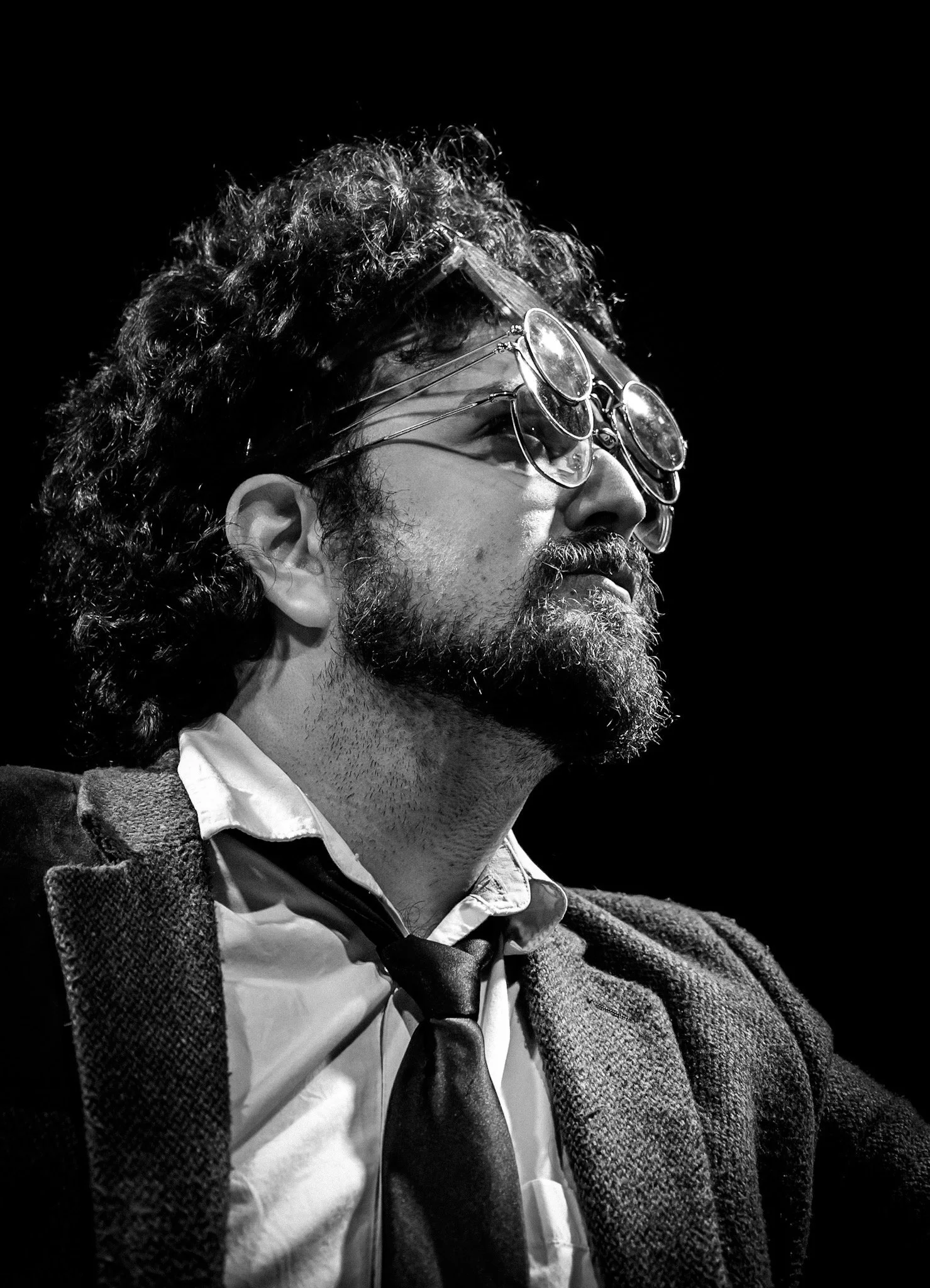 Black and white photo of a man with curly hair and glasses, wearing a suit jacket, shirt, and tie, looking to the right against a dark background.
