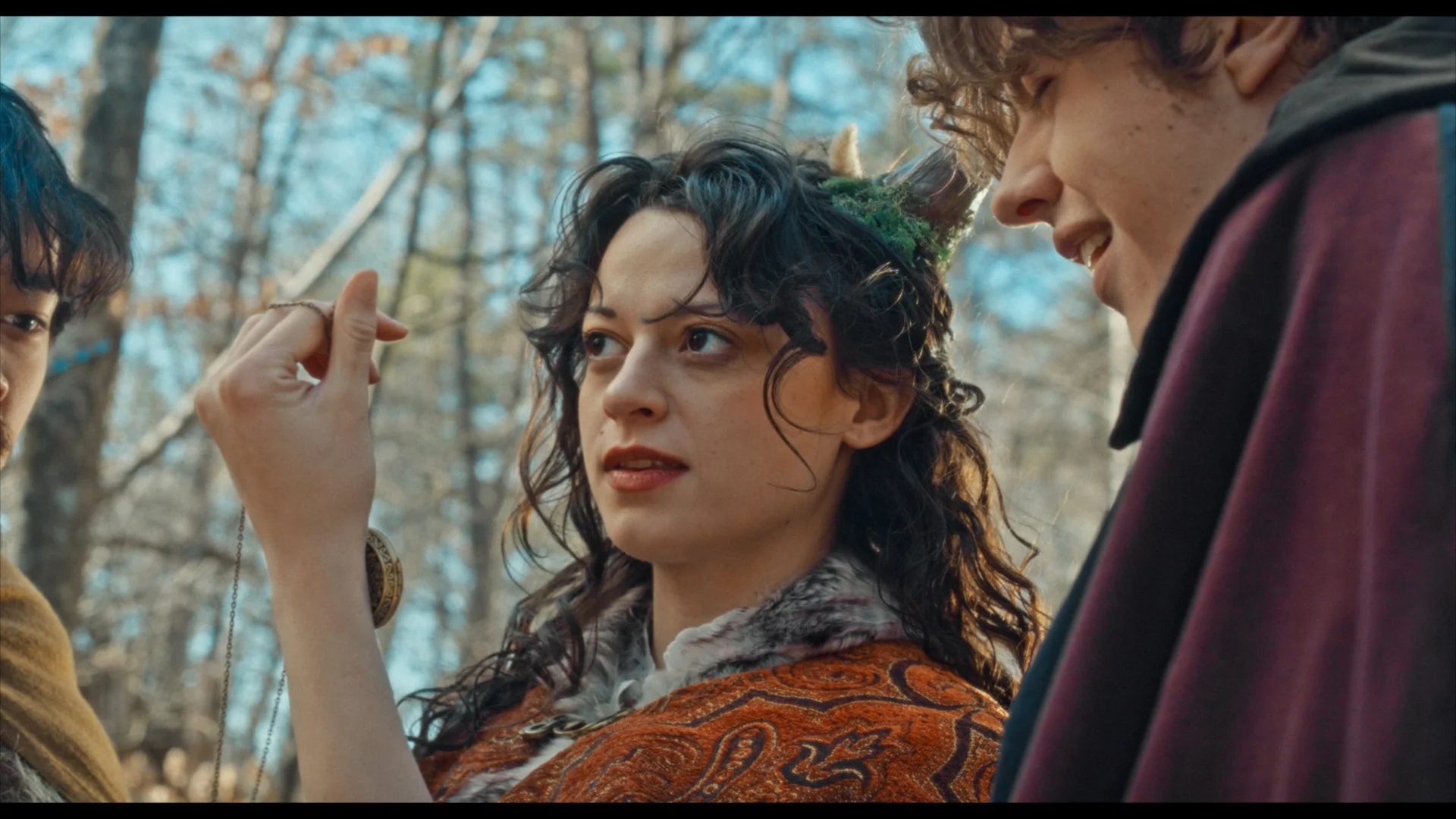 Three people outdoors, with trees and bare branches in the background. The woman in the center, with curly dark hair and a green moss hat, is looking at the man on the right. The man on the right, with curly blond hair, is smiling and looking at the woman. The person's hand on the left is holding a pendant or keychain near the woman’s face.